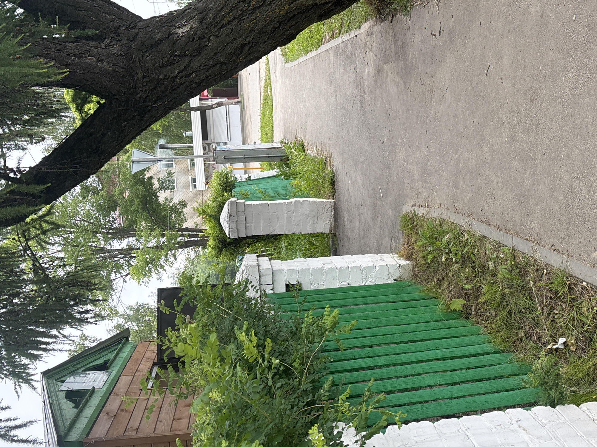 The image displays an outdoor street scene in Красные Баки, Russia, under bright daylight. A wide asphalt or concrete pedestrian path runs diagonally from the lower right foreground towards the upper left background. To the left of this path, a fence composed of white brick pillars and horizontal green wooden slats is prominently featured, extending from the foreground into the midground.
Lush green bushes and foliage grow abundantly both in front of and behind the fence, with some grass and weeds visible along the edge of the path. A large tree trunk, featuring rough, dark bark, stretches diagonally across the upper left portion of the frame, partially obscuring the background.
Beyond the fence and vegetation, several buildings are visible in the midground. On the far left, there is a rustic wooden building with a pitched green roof that appears to have some silver-colored repairs or patches. Further right, a light-colored brick building with windows is present, along with another structure that has a roll-down security shutter, possibly indicating a commercial establishment. A metallic triangular sign, likely a road or informational sign, stands near these buildings. Utility poles and wires are also discernible in the distance.
No people are present in the scene, which conveys a quiet, static atmosphere of a residential or mixed-use area. Some indistinct Cyrillic text, possibly including the letters "АРА," is visible on a sign affixed to one of the buildings in the midground. The weather appears to be clear or partly cloudy, with no indication of rain.