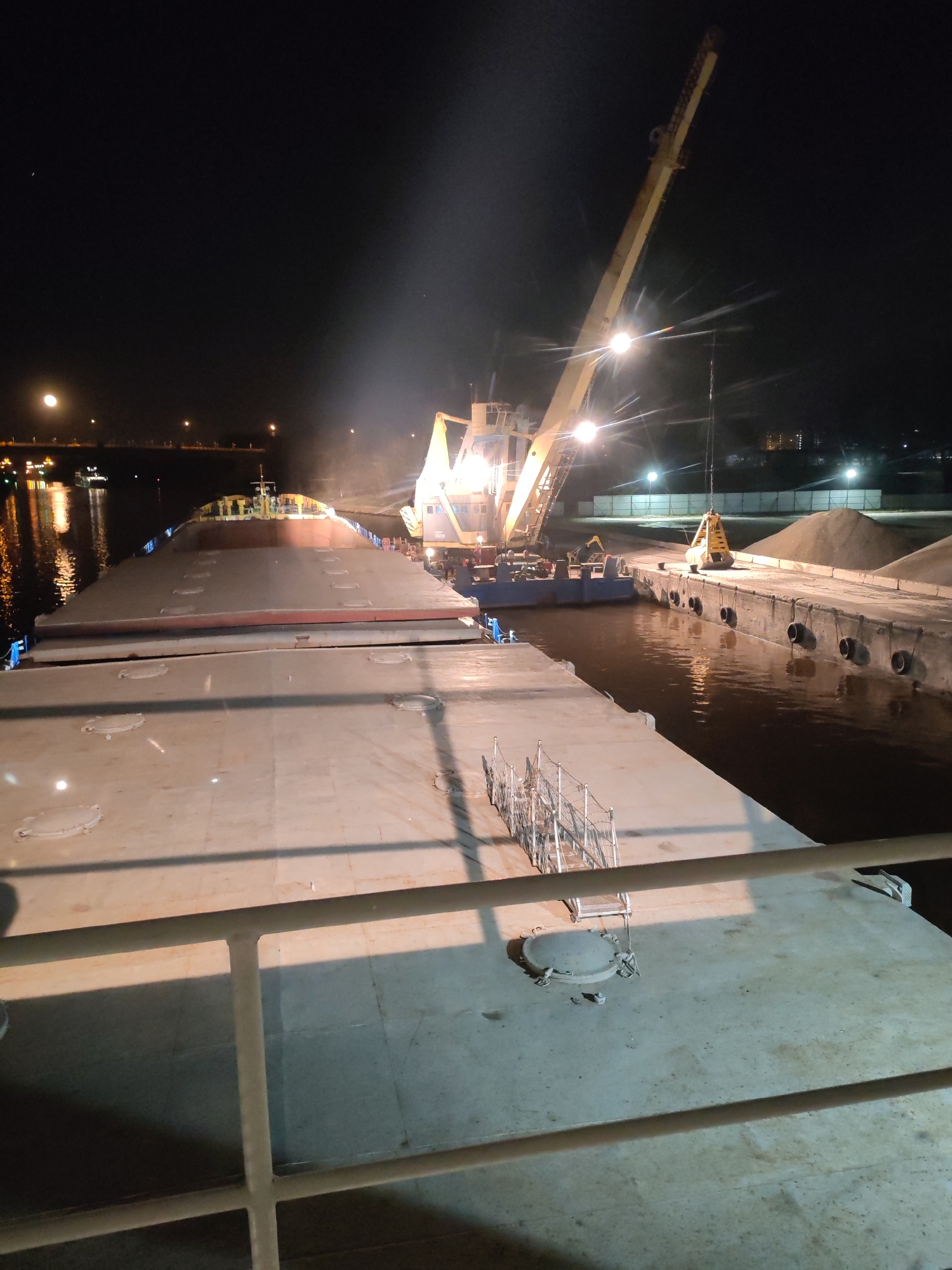 The image depicts a nighttime scene on a waterway in what is likely Yakhroma, Russia. A large barge, presumably used for transporting materials, dominates the foreground, its deck featuring several round hatches and a ladder structure. To the right of the barge, a crane is actively engaged in loading or unloading materials from the vessel onto a dock. The dock is lined with rubber bumpers and has mounds of material stockpiled on it. Beyond the dock, there are buildings and trees silhouetted against the night sky, illuminated by distant lights. The waterway reflects the lights from the dock, crane, and surrounding area. There are no people visible in the image. The scene is well-lit by artificial lighting, providing a clear view of the industrial activity taking place. The crane appears to be in operation, its arm extended and equipped with a grab bucket.