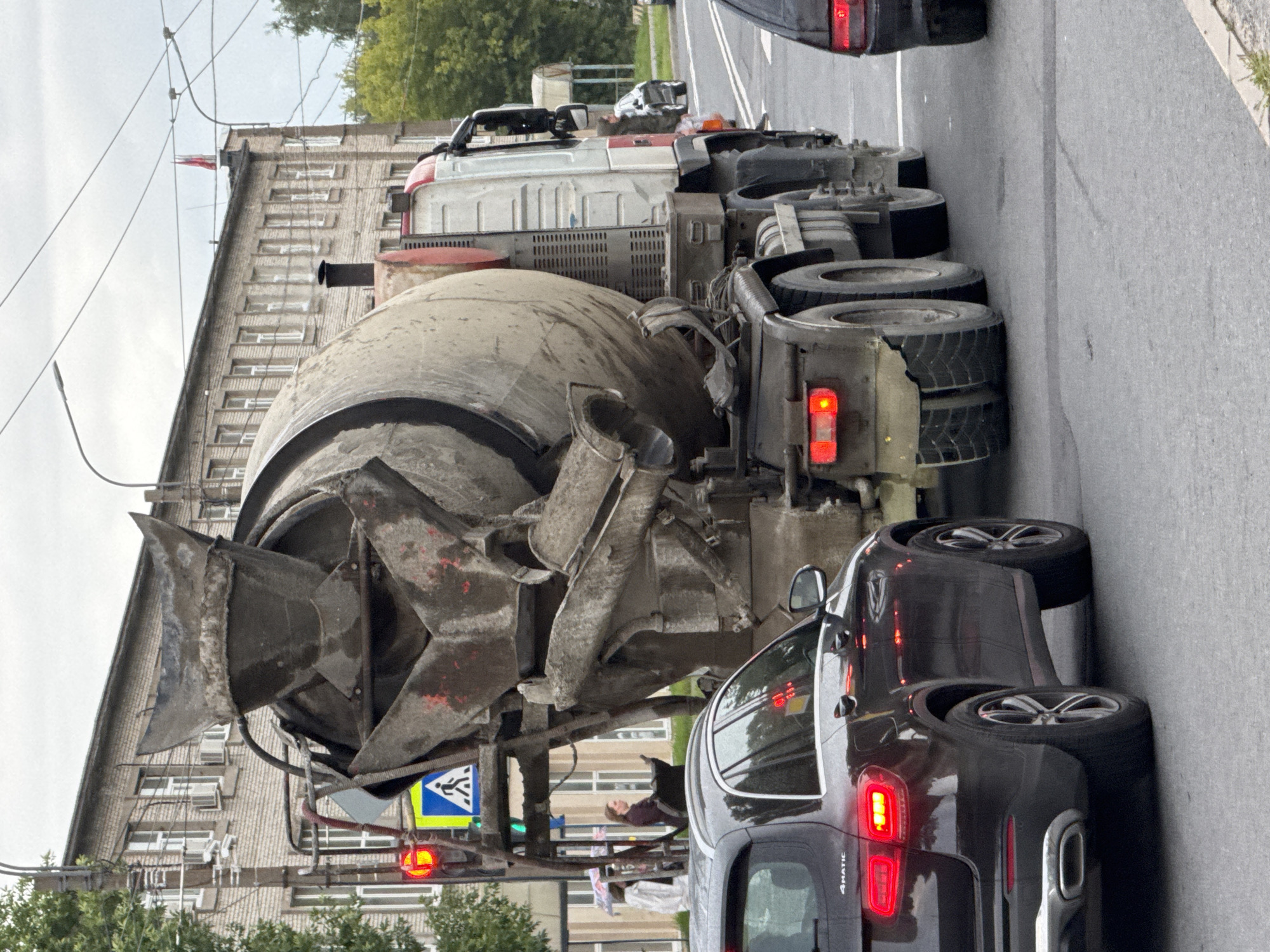 The image shows a concrete mixer truck driving on a street in Saint Petersburg, Russia, with a dark-colored SUV in front of it and another vehicle partially visible behind the truck. The concrete mixer truck is large and appears to be made of concrete with red accents on the cabin. It has a rotating drum filled with cement. The SUV is a Mercedes-Benz GLC, indicated by the "4MATIC" badge on its rear. Both vehicles are on a paved road, and a building with multiple windows and a brick facade is visible in the background, suggesting an urban environment. A traffic light is showing red, and a pedestrian crossing sign is also present. The sky is overcast. Two people are visible near the traffic light, one holding an umbrella. The scene appears to be a street in an industrial or residential area of the city.