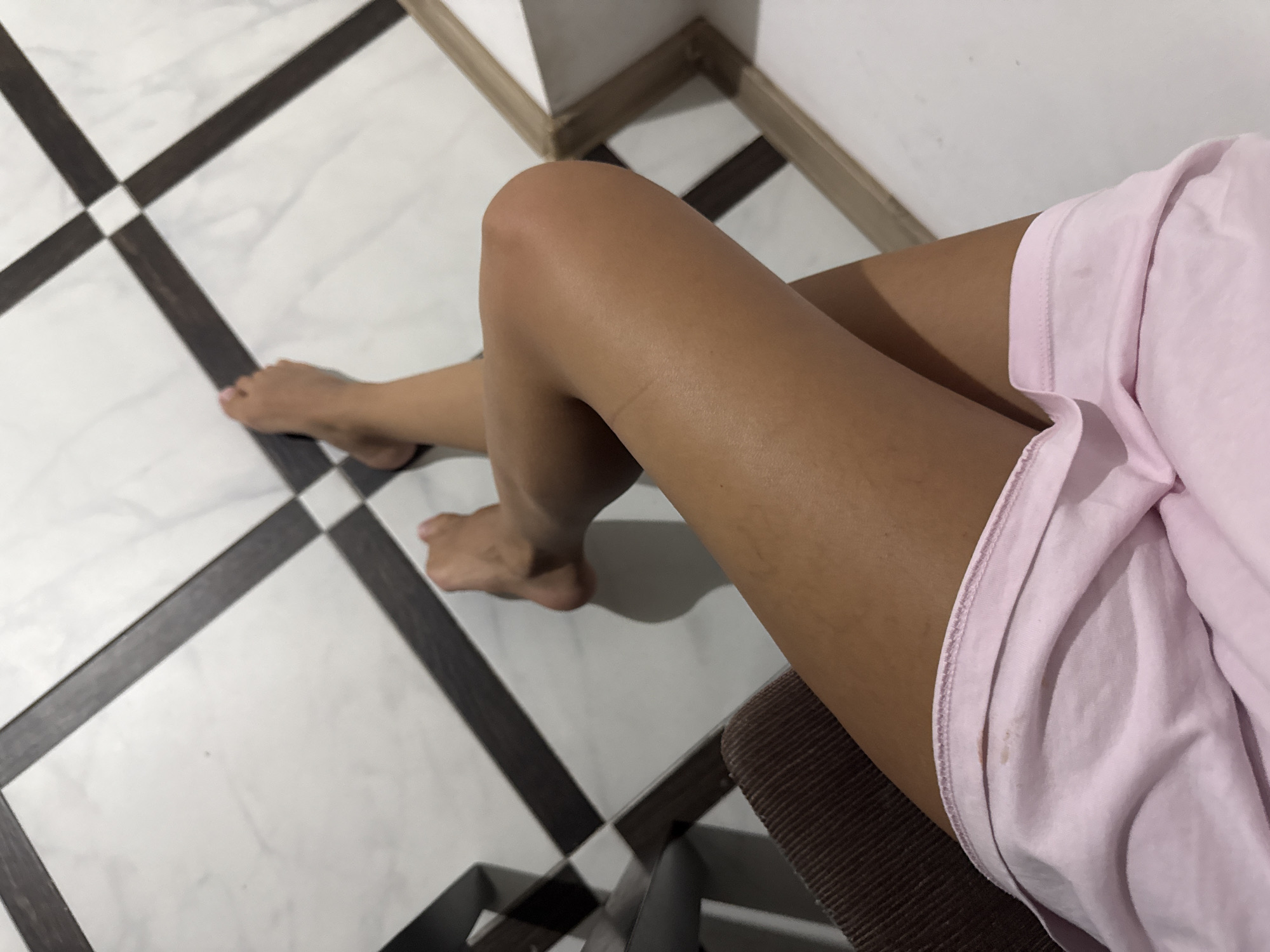The image shows a person's legs and feet resting on a tiled floor. The tiles are white with dark brown square accents forming a diagonal pattern. The person is wearing pink shorts. One leg is crossed over the other, and both feet are visible. The lighting suggests an indoor environment, possibly a room or hallway. There is no specific information in the image to indicate a location in Chișinău, Moldova.