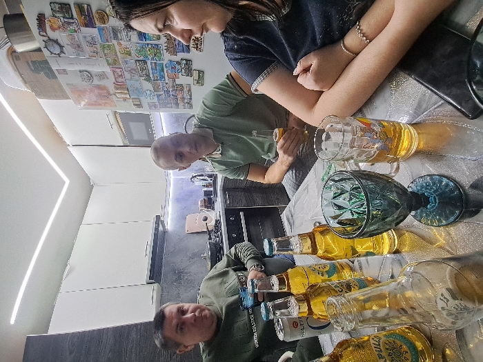 A group of people are gathered around a table in a kitchen. There are bottles of beer and filled glasses on the table. One person is pouring beer into a glass. The kitchen has white cabinets and a modern design. There are magnets and other items attached to the refrigerator.