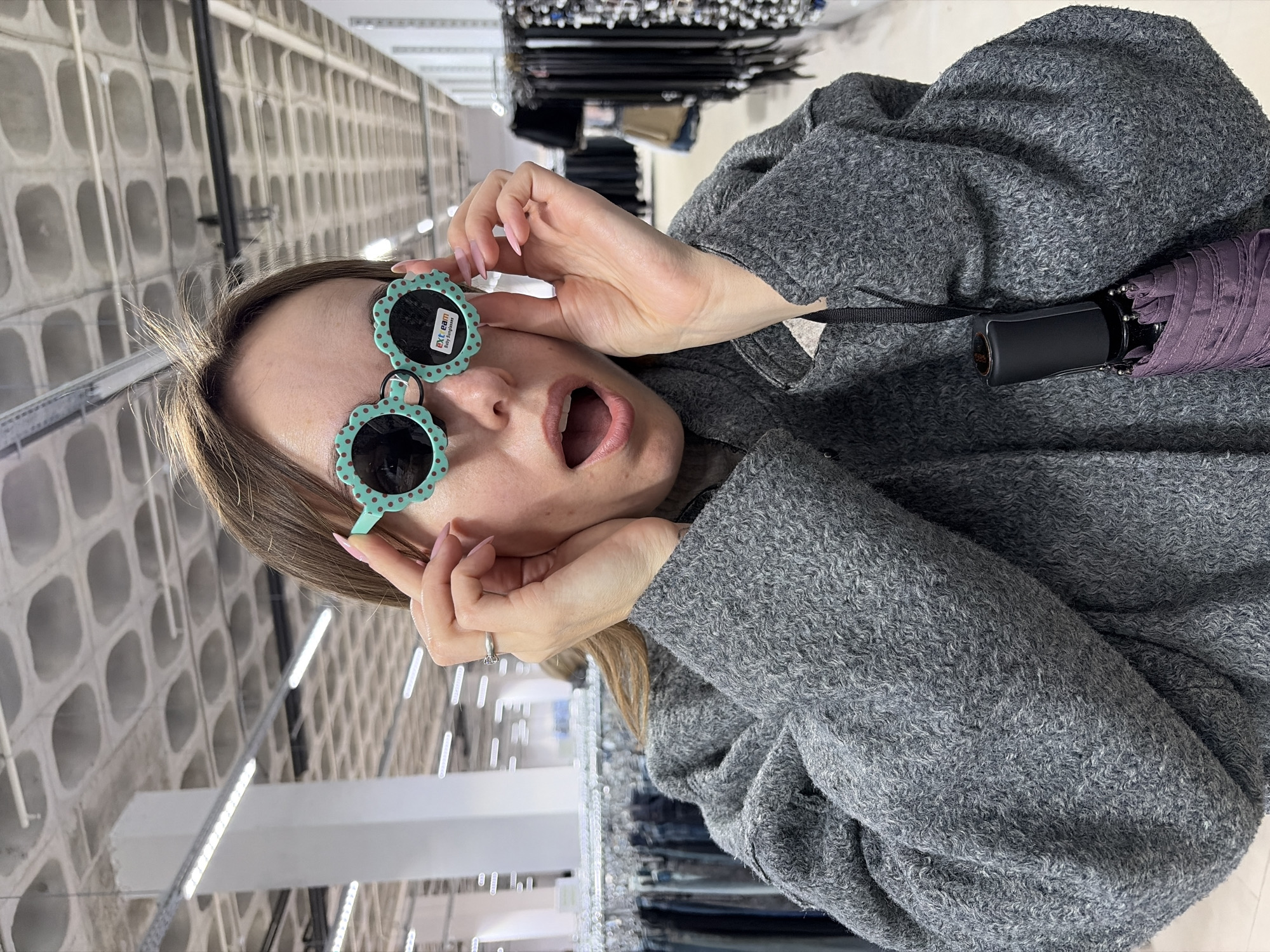 A woman with light brown hair is shown indoors, likely within a retail store or warehouse, indicated by the rows of shelving units and concrete block-like structures in the background. She is wearing a grey, textured coat. Her hands, featuring long light pink nails and a ring on her left ring finger, are holding a pair of distinctive turquoise, flower-shaped sunglasses with brown polka dots up to her eyes. Her mouth is open in an expression of surprise or playful awe.
Tucked into her coat on the right side of the image is a small, folded purple umbrella. The background also shows bright linear fluorescent lights and clothing racks with garments. Text visible on the lens of the sunglasses reads "EYE eam" and "Baby Sunglasses," accompanied by a small, indistinct logo. The overall scene captures a candid moment of the woman trying on or reacting to the sunglasses in a functional, commercial setting.