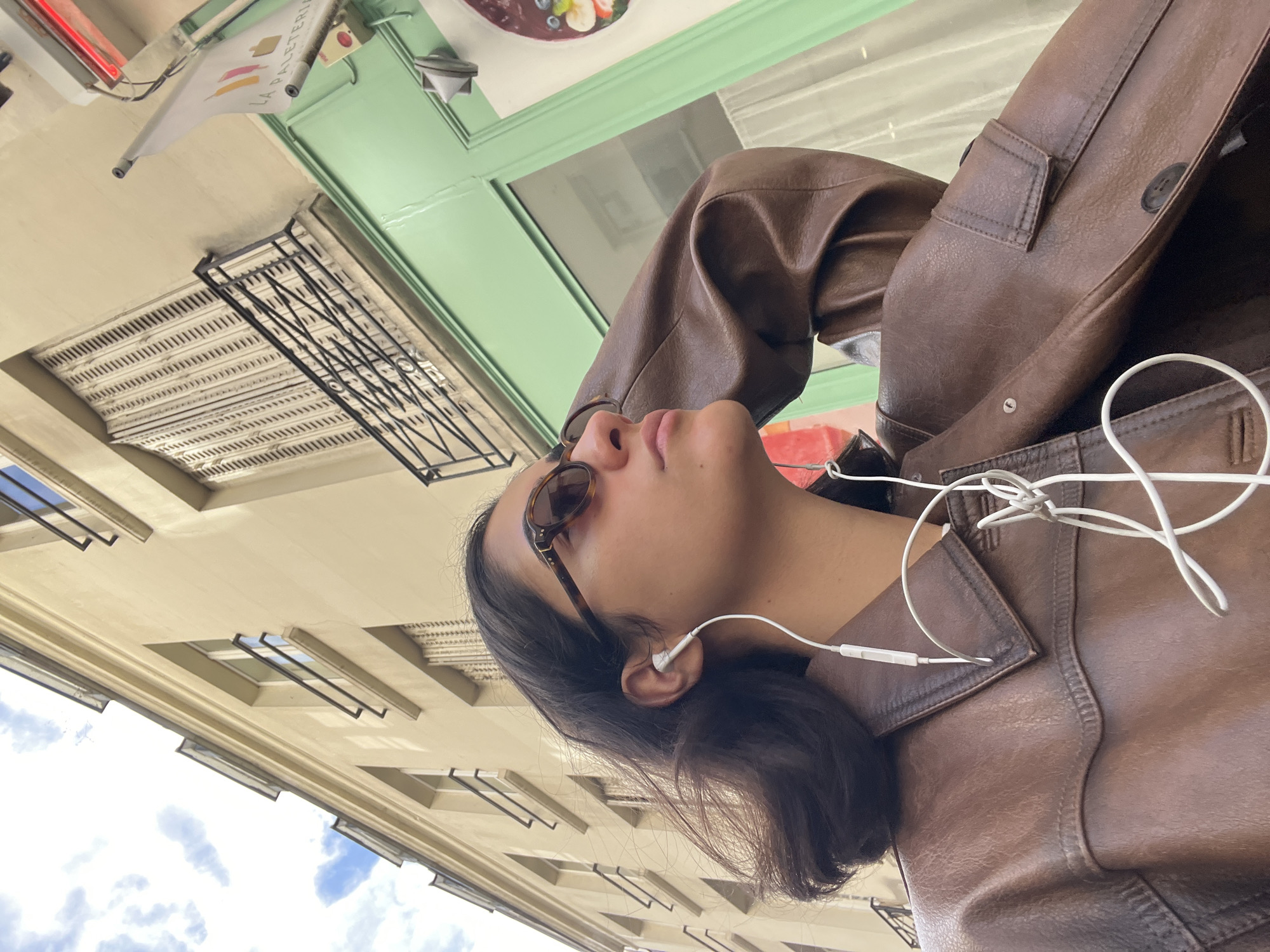 The image is an upward-angled shot capturing a person and the architectural details of a building in Paris during the daytime.
**Subjects:** The primary subject is a young individual with dark hair, seen in profile, looking upwards. They are wearing a brown leather jacket and round tortoiseshell-patterned sunglasses. White wired earbuds are visible, inserted into their ears. The background features a multi-story building.
**Activity or event:** The person appears to be walking casually along a street, listening to music or a podcast, and looking up, possibly observing the architecture or the sky.
**Notable details:**
* **Time of day:** It is daytime, indicated by the bright, even lighting and the blue sky with white clouds visible above the buildings.
* **Weather:** The weather appears fair and sunny with some cloud cover.
* **Location cues:** The classic European architectural style of the building, with its ornate facade, decorative grates, and distinctive windows, strongly suggests an urban setting, consistent with Paris.
* **Emotions:** The person's expression is neutral and contemplative, simply looking upwards.
* **Visible text:** A white banner or flag is visible near a green awning, bearing the text "LA PALETERIA" in a colorful, stylized font. This likely indicates the name of a shop or business.
The overall scene captures a candid moment of an individual enjoying a walk through a city, immersed in their surroundings and music.