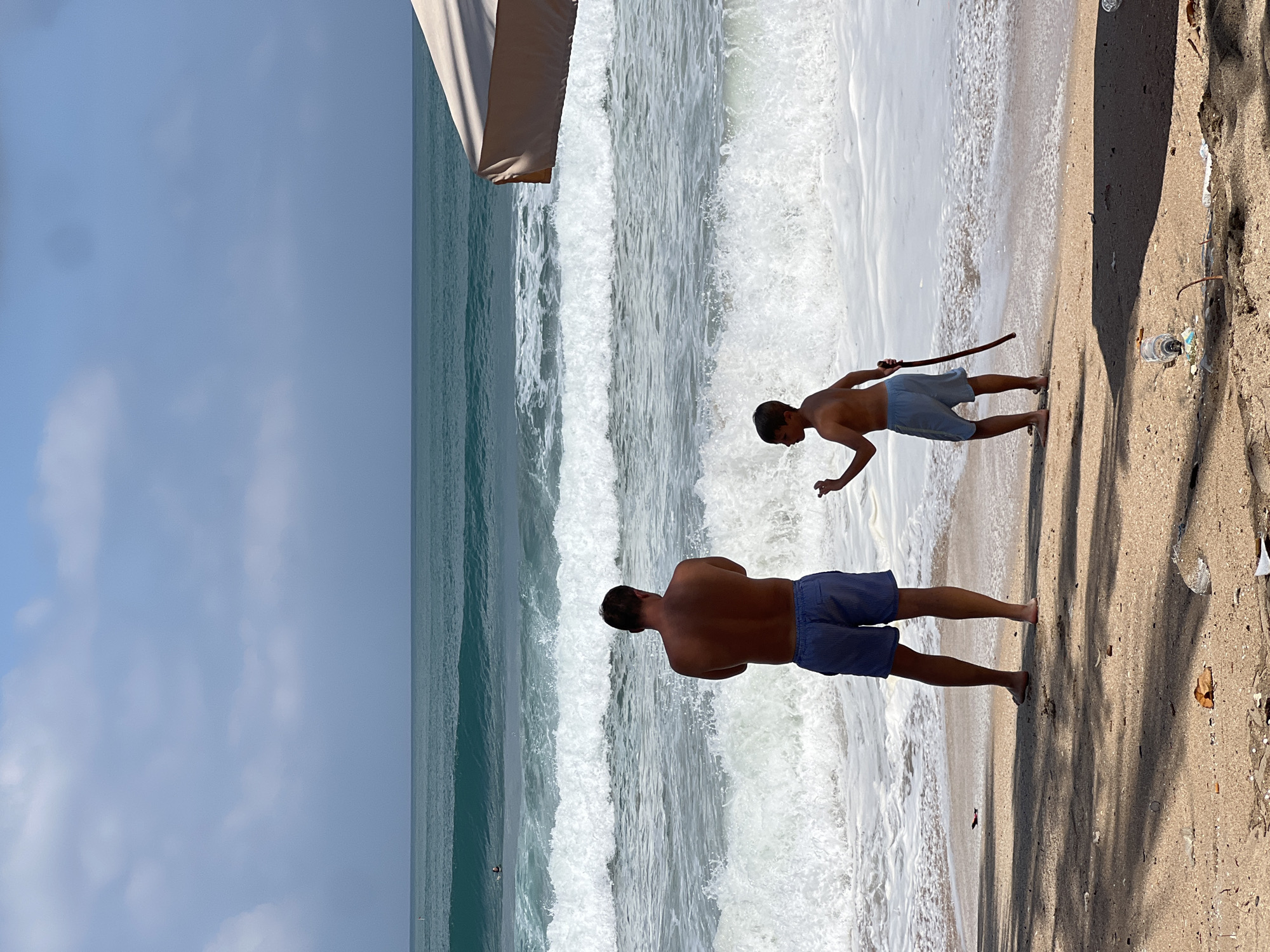 A man and a boy stand on a sandy beach in Kuta, Indonesia, facing the ocean. The man, from the back, wears blue patterned shorts. The boy, also facing the ocean, wears light blue shorts and holds a stick. The ocean has large waves crashing on the shore, and the sky is partly cloudy. In the background, a surfer is visible in the distance. The beach is littered with some trash. A portion of a tan umbrella is visible on the right side of the frame.