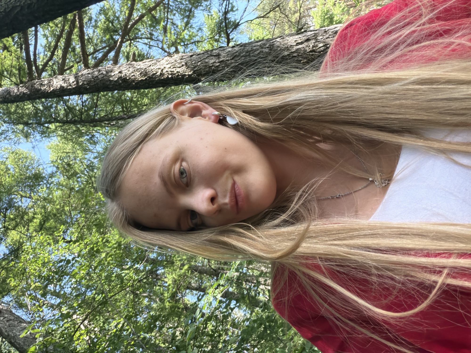 This is a head and shoulders selfie of a young woman with long, blonde hair, taken outdoors in a forest. She is wearing a red top over a white shirt and a silver necklace with a pendant. She has a single earring in her left ear. The background is filled with green foliage and tall trees, with patches of blue sky visible. The lighting suggests it is daytime. There are no identifiable landmarks that would place this scene specifically in Izhevsk, Russia.