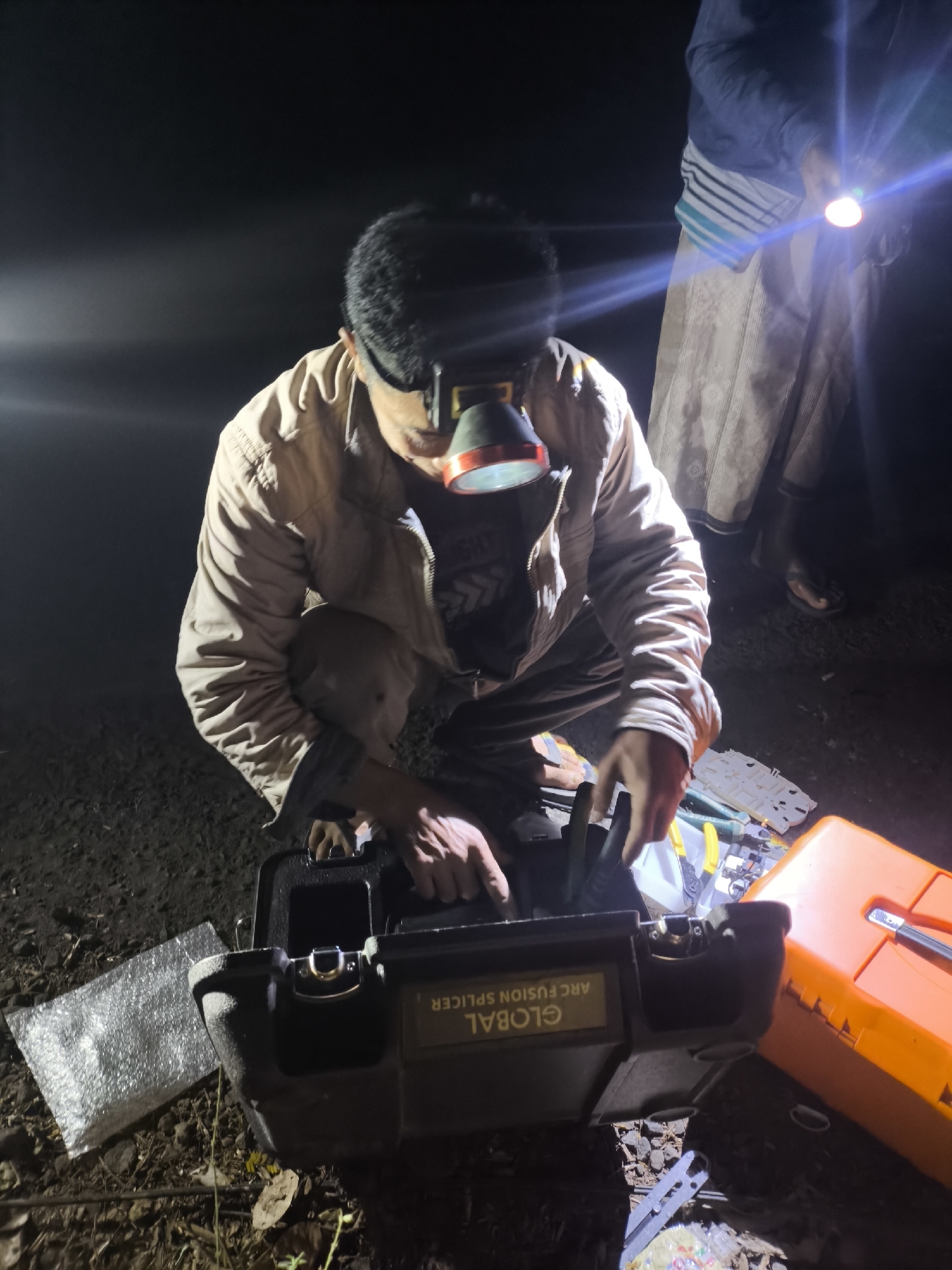 In the city of Bodang, Indonesia, at night, a person wearing a headlamp is kneeling and working on a black toolbox. The toolbox has "GLOBAL ARC FUSION SPLICER" written on it. Tools, including wire cutters and a white component, are spread out on the ground next to the toolbox, along with a sheet of bubble wrap. An orange toolbox is also present. Another person, holding a flashlight, stands to the right, illuminating the scene. The ground is covered in dirt and debris.