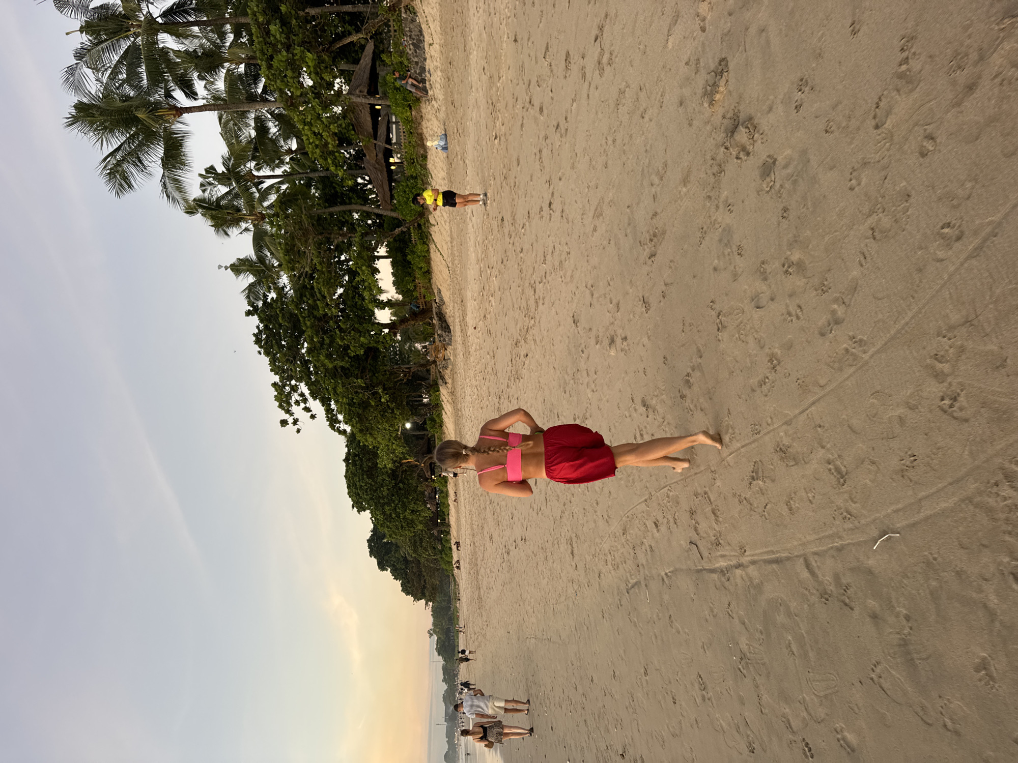 The image shows a beach scene in Kuta Selatan, Indonesia. The foreground is sandy, with footprints visible in the sand. A woman wearing a pink top and red skirt is walking barefoot on the beach. In the background, other people are visible on the beach. A line of trees, including palm trees, borders the beach. The sky is light, suggesting either early morning or late afternoon.