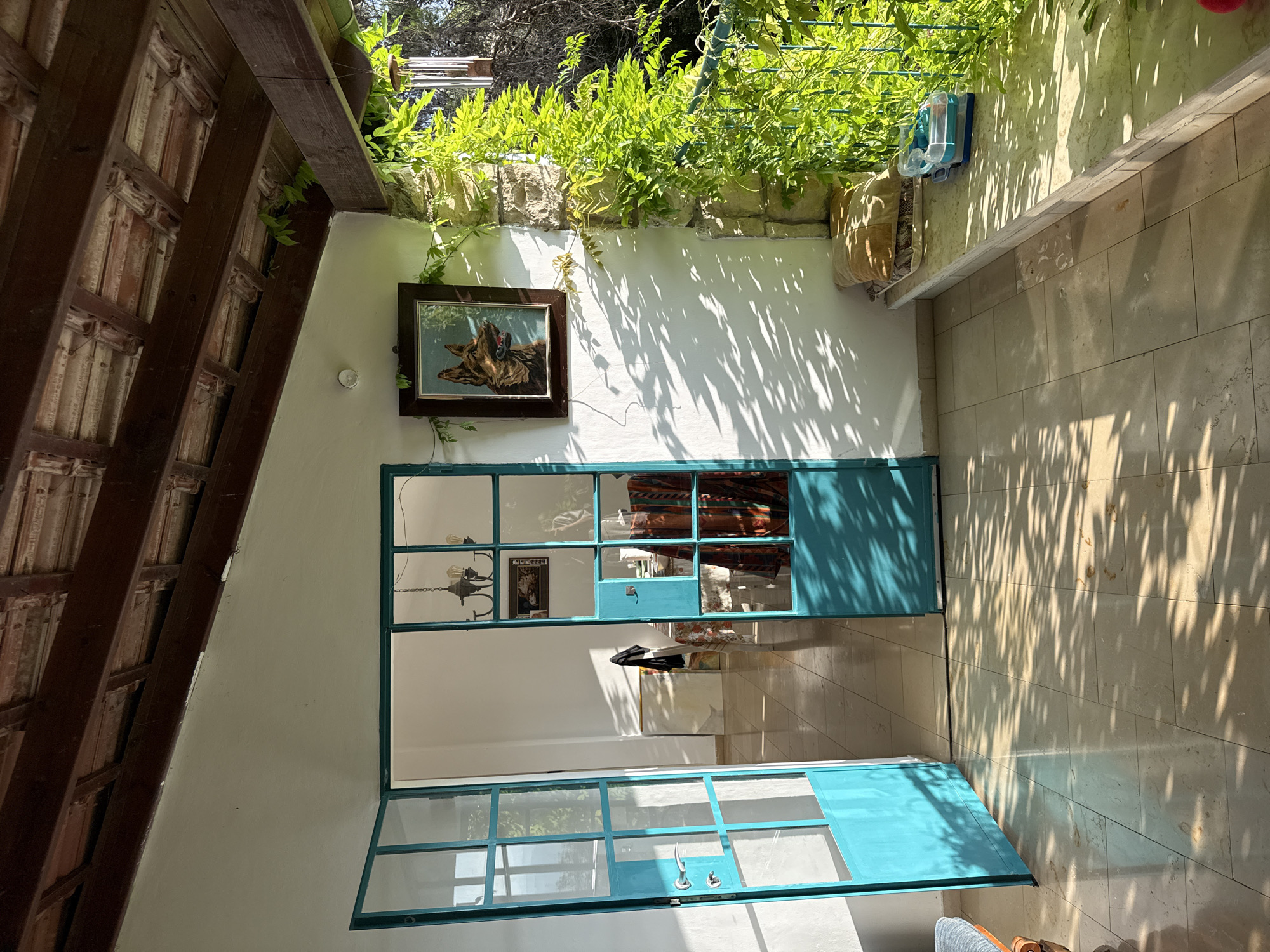 The image depicts a sun-drenched patio area with a covered roof in Kfar Vradim, Israel. Two teal-colored glass doors are open, revealing an interior space with tiled floors and white walls. Sunlight casts dappled shadows from foliage onto the patio floor and walls.
On the patio, a stone ledge runs along the right side, adorned with two cushions and a blue and clear plastic container. Lush green vines climb the stone pillar and hang over the ledge, suggesting a garden setting. A wind chime is visible hanging from the wooden roof structure.
The interior visible through the doors features tiled floors, white walls, and a chandelier. A framed picture of a dog hangs on the exterior wall, to the left of the right door. The overall impression is one of a relaxed, outdoor living space bathed in natural light.