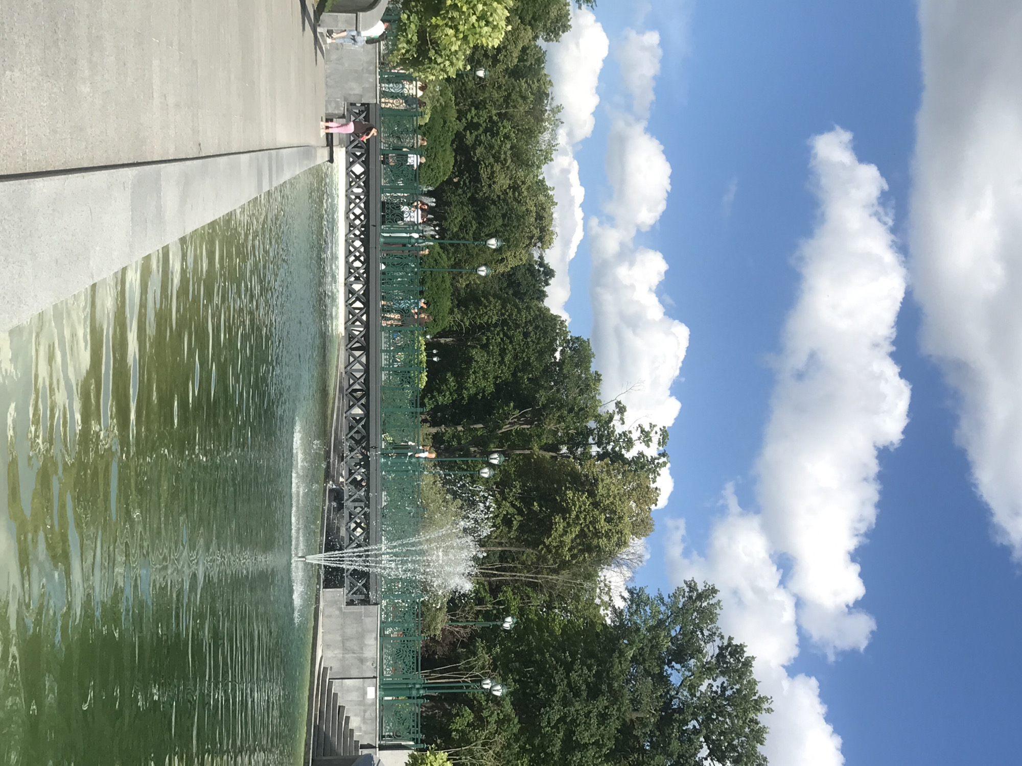 The image depicts a sunny day in a park with a wide, calm body of water, likely a pond or canal, in the foreground. A multi-tiered fountain sprays water in the center of the pond. A grey concrete walkway runs along the left side of the pond, with a few people visible. Across the pond, a dark-colored bridge with decorative metal railings spans the water, and several people are walking on it. Behind the bridge, a dense line of green trees forms the backdrop, with a clear blue sky dotted with white clouds above. Street lamps with white lights are visible along the bridge and the treeline. Based on visual cues, this scene is identifiable as a section of the Shevchenko Garden (Sad imeni Tarasa Shevchenka) in Kharkiv, Ukraine.