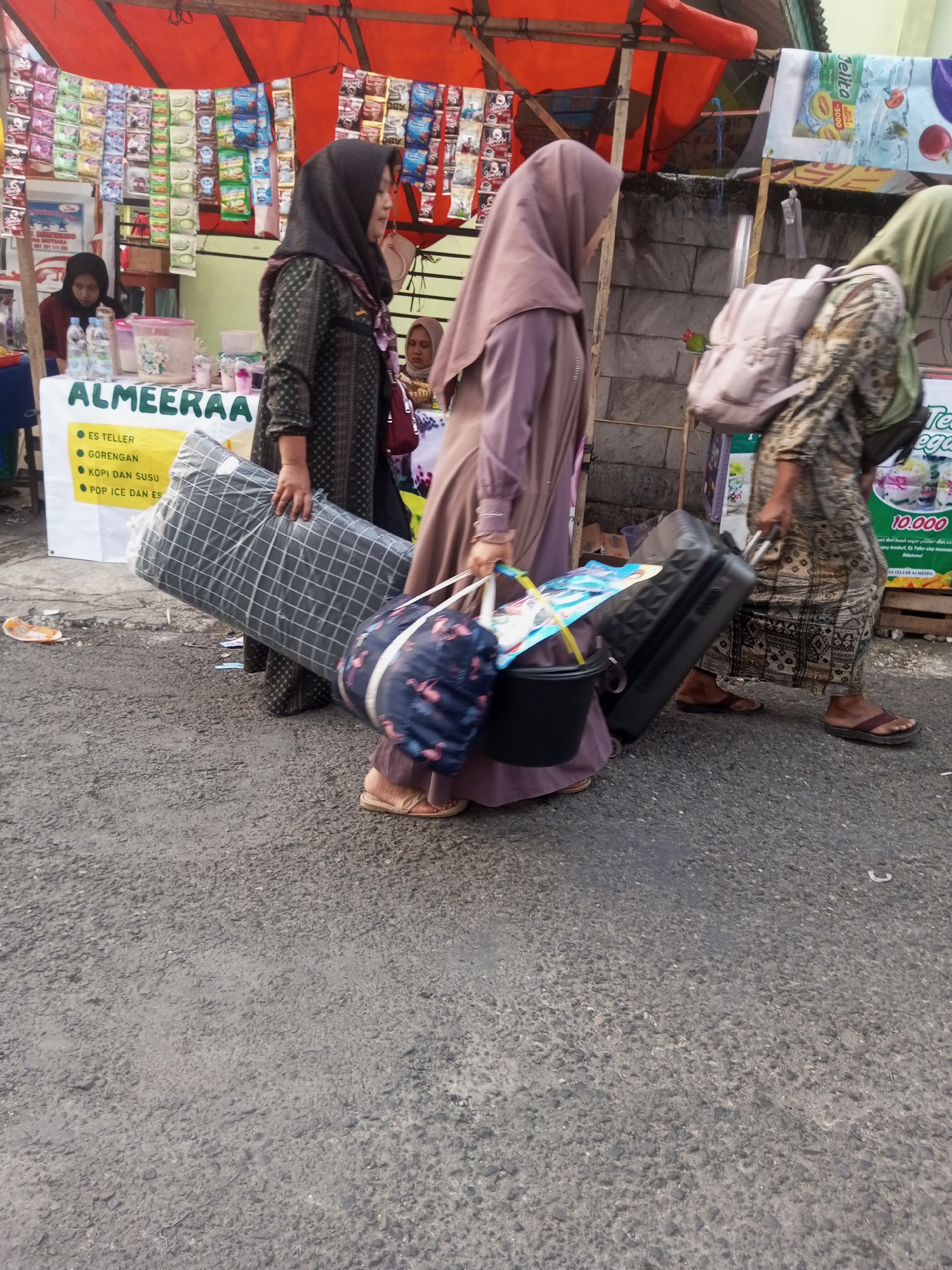 A group of people are walking on a street in Pandansari, Indonesia, carrying various items. A woman wearing a hijab and a long patterned dress walks in the foreground, pulling a black suitcase with a patterned bag on top of it. To her left, another woman wearing a hijab and a patterned brown and white dress carries a large, rolled-up item wrapped in plastic. Behind them, a woman in a light brown hijab walks away from the camera, wearing a backpack. In the background, a market stall displays various packaged goods. The ground is paved with asphalt.