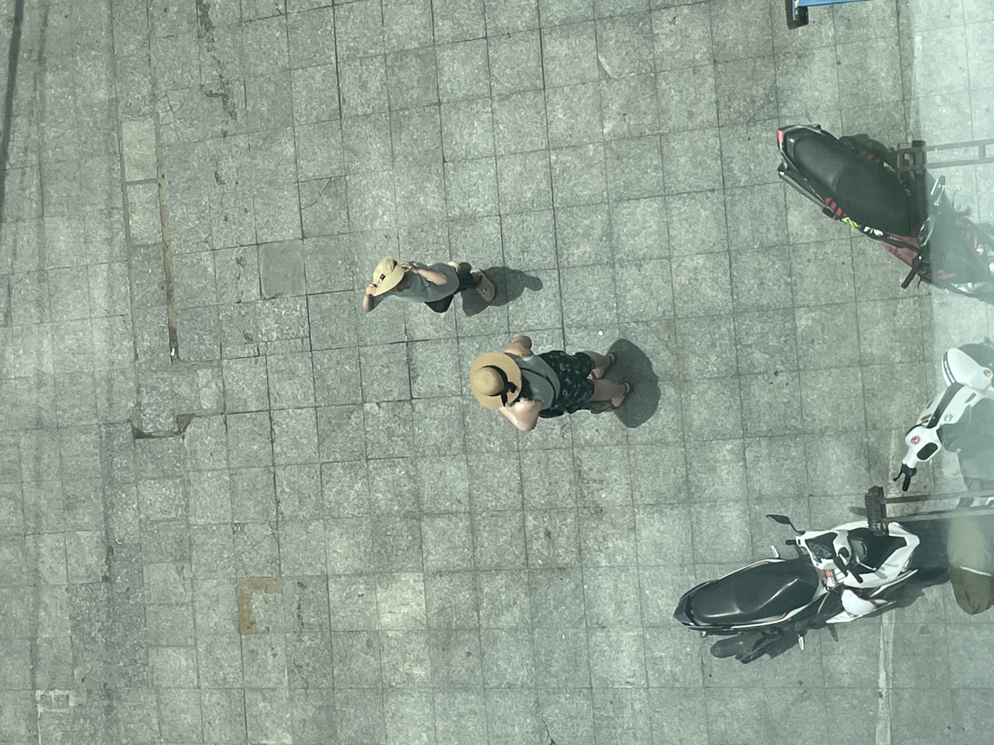 The image shows an overhead view of two individuals on a paved city square in Nha Trang, Vietnam. Both individuals are wearing wide-brimmed straw hats, suggesting a sunny day. One person is wearing a light-colored t-shirt and patterned shorts, and appears to be standing with their hands behind their head. The other person is wearing a t-shirt and dark shorts, and is holding their hat in their hand. Two motorbikes are parked on the paved area. The ground is made of square tiles. The overall scene is a snapshot of everyday life in a public outdoor space.