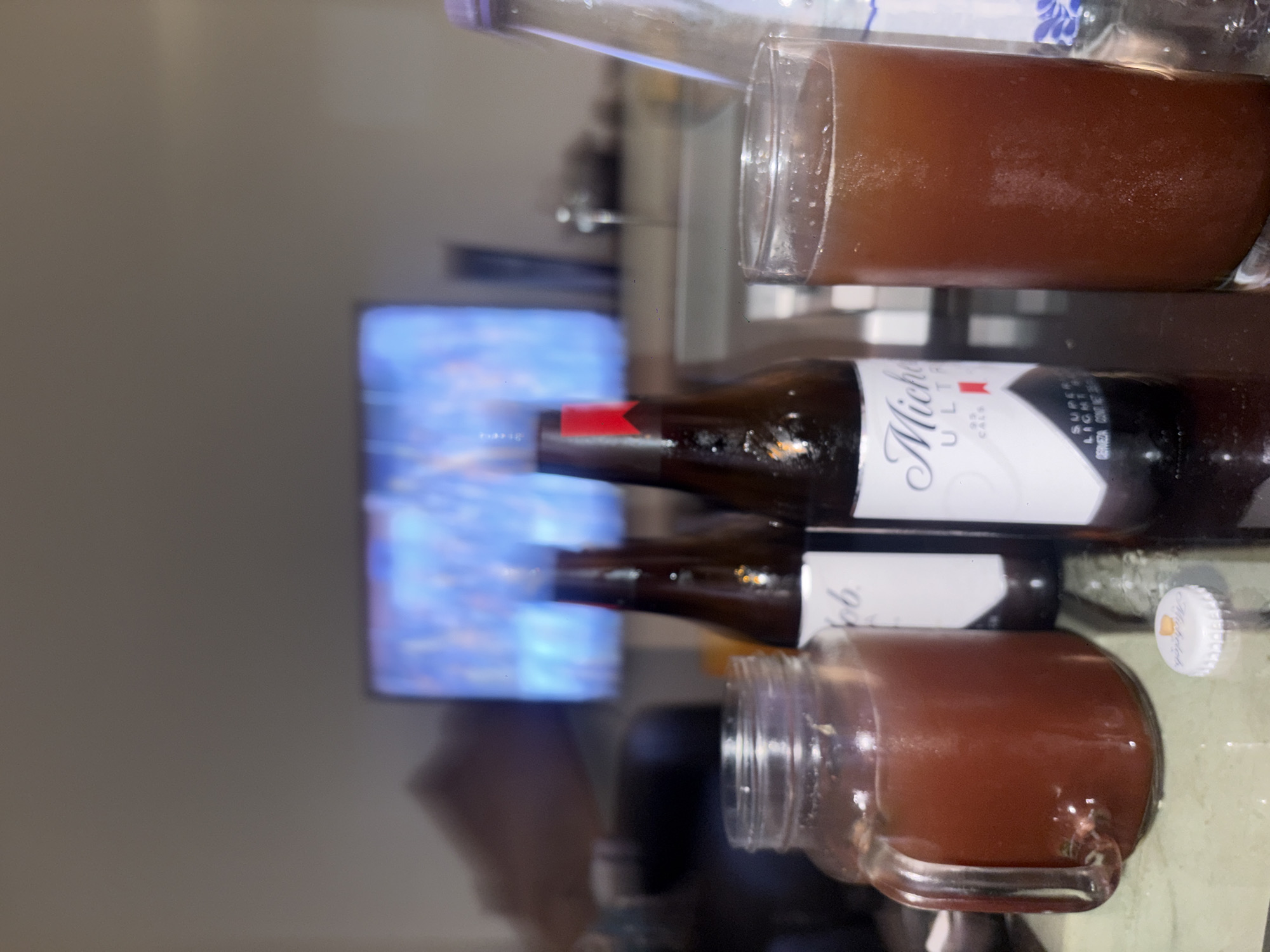 The image depicts a tabletop scene with drinks and beer bottles. The scene appears to be indoors, possibly in a home in Mexico City, Mexico, as suggested by a bottle of Michelob Ultra beer, a popular brand in Mexico. The lighting is soft and ambient, suggesting a relaxed atmosphere.
On the table, there are two Michelob Ultra beer bottles, one seemingly full and the other potentially empty or partially consumed. There are also two glasses filled with a brown liquid, which could be beer, mixed drinks, or another beverage. One glass is a regular pint glass, while the other is a mason jar with a handle. A plastic bottle with a light purple lid is visible behind the beer bottles and a pint glass. There is a white beer bottle cap laying on the table behind the jar and beer bottles.
The surface the items are on is reflective, suggesting a polished table or countertop. In the background, a television or monitor displaying blue tones is visible, indicating the setting is a place where entertainment is available. The overall composition gives the impression of a casual social gathering or a quiet evening at home.