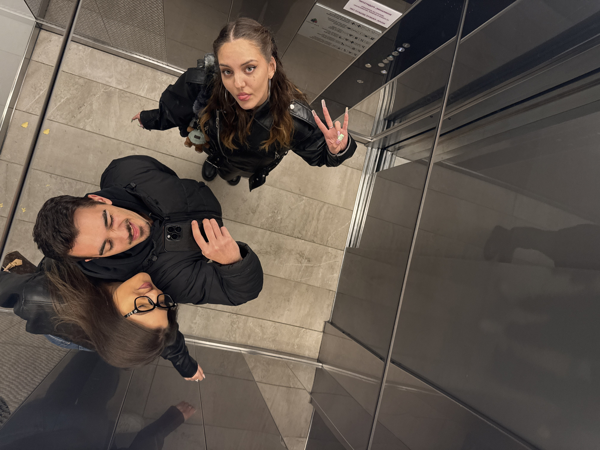 Two women and one man are in an elevator. The man is holding a cell phone. The woman at the top of the frame is throwing the "sign of the horns." The elevator is in Sofia, Bulgaria.