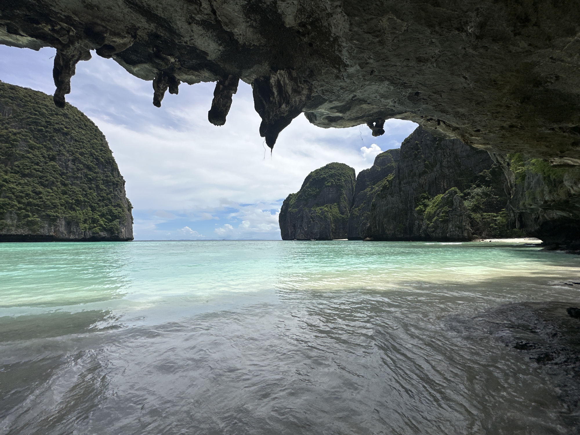 The image captures a view from within a cave opening, looking out onto a tropical bay in Ao Nang, Thailand. The foreground is dominated by the dark, rough texture of the cave ceiling, with stalactites hanging down. Beyond the cave's mouth, the scene opens to reveal a calm, turquoise and aquamarine bay. Steep, jungle-covered limestone karsts rise dramatically from the water on either side of the bay, framing the view. The water is crystal clear, with a sandy bottom visible in the shallower areas. The sky is partly cloudy, with soft white clouds scattered across a pale blue expanse. The overall impression is one of natural beauty and serene isolation. There are no people or man-made objects visible in the image.