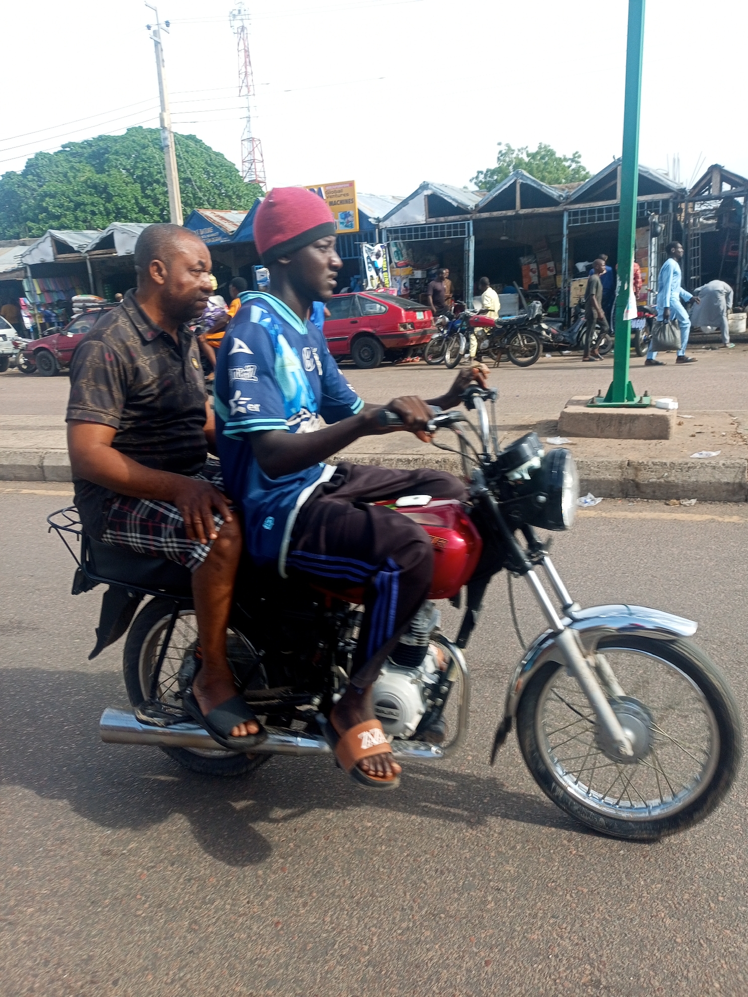 The image depicts a daytime street scene in the city of Unknown, Unknown, featuring two males riding a red motorcycle. The rider, a younger male, wears a red beanie, a blue and white short-sleeved jersey, dark trousers with blue side stripes, and tan slip-on sandals, gripping the handlebars and facing forward. The passenger, an older male, sits behind him, wearing a dark patterned polo shirt, plaid shorts, and dark slip-on sandals. The motorcycle features a red fuel tank, black seat, chrome front fender and exhaust, and a black headlight with white material around its base. The visible engine casing indicates "BAJAJ."
The motorcycle is on a paved road, with a curb and sidewalk visible to its right. In the background, a row of single-story market stalls or shops with metal roofs lines the street. One shop displays a yellow sign for "GLOBAL VENTURES MACHINES." Several pedestrians are visible around the shops, including a man in a light blue garment walking left with a dark bag and another person bending over near a container. Various vehicles, including a red sedan and parked motorcycles, are also present. Utility infrastructure includes a wooden pole with wires on the left, a tall green pole on the right, and a distant red and white lattice telecommunications tower, with a large green tree behind the shops on the left. Shadows indicate bright sunlight.