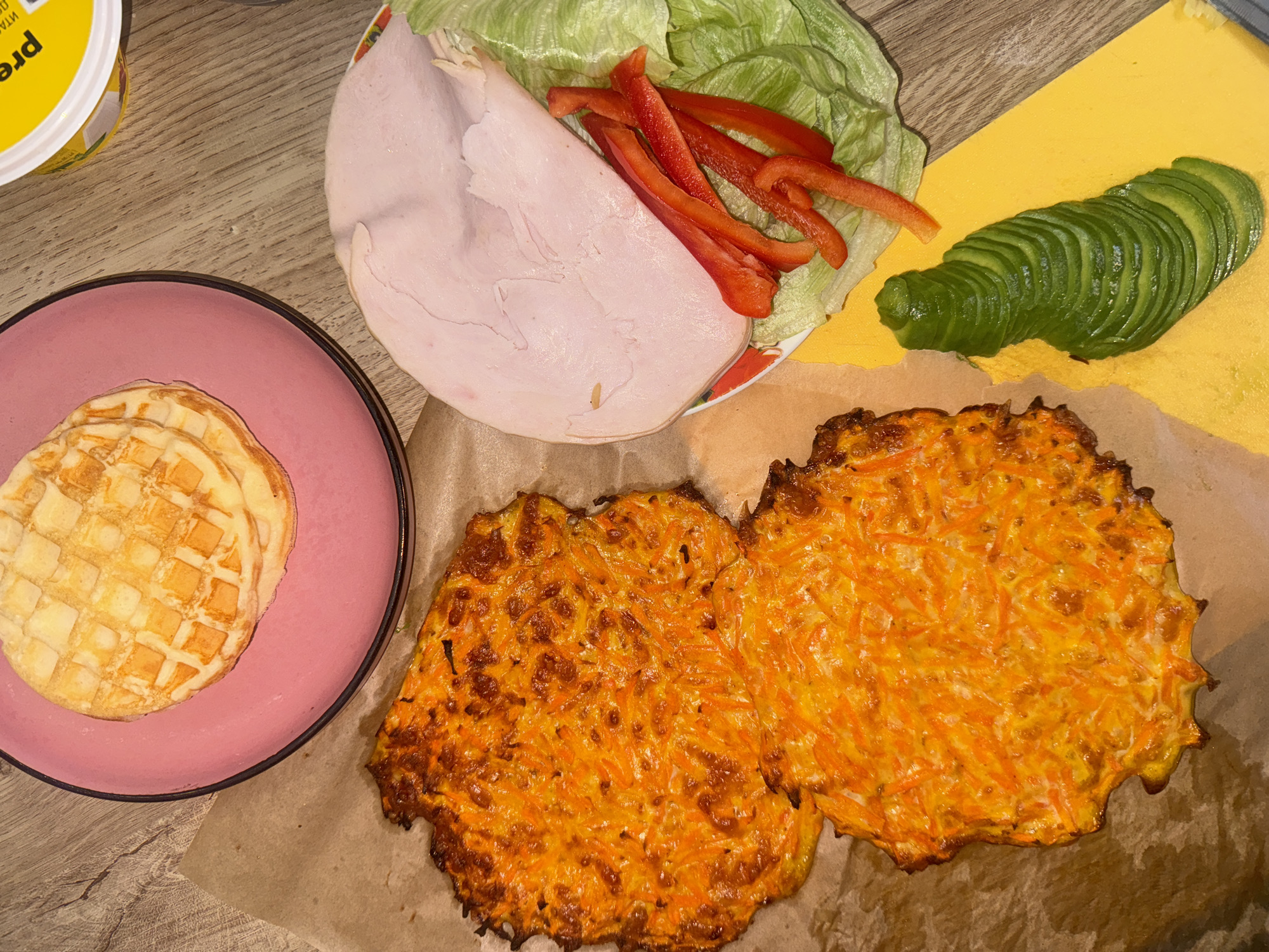 The image shows a table with various food items, seemingly prepared for a meal. On a pink plate, there are three waffles. Next to it, there are two orange-colored, irregularly shaped items that appear to be shredded carrot fritters on brown paper. A plate contains lettuce, slices of red bell pepper, and a large slice of turkey breast. Sliced avocado lies on a yellow surface. A yellow container is partially visible in the upper left corner. The table surface is light brown with a wood grain pattern. There is no indication of the location of Krasnoyarsk, Russia.