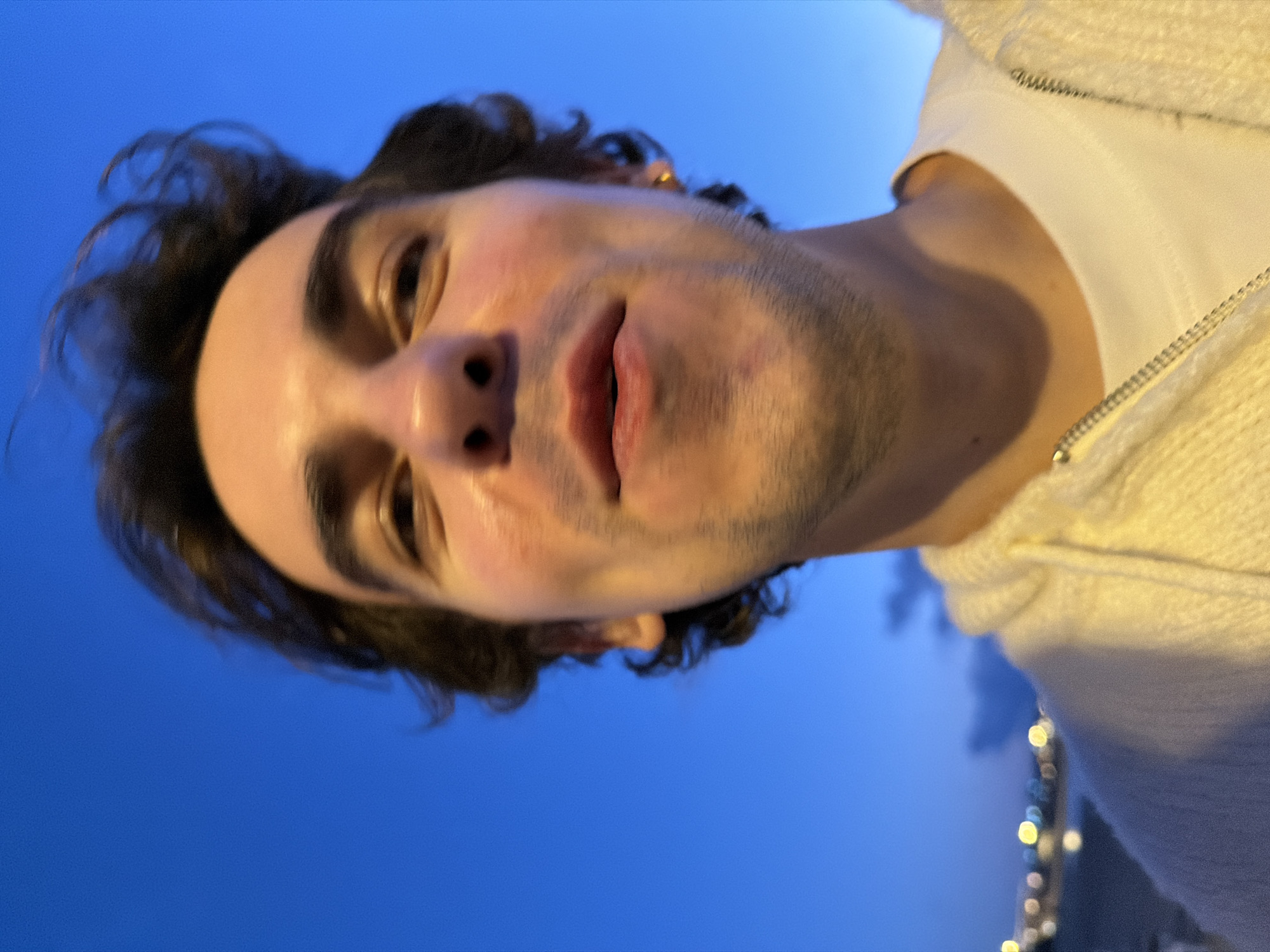 The image is a close-up, tilted selfie of a young man with dark hair and stubble. He is wearing a white t-shirt and a light-colored, possibly cream-colored, zip-up hoodie. The background is a blue sky. At the bottom right corner, blurred lights suggest the presence of a cityscape. Based on the image's metadata, the location is likely Tel-Aviv, Israel.