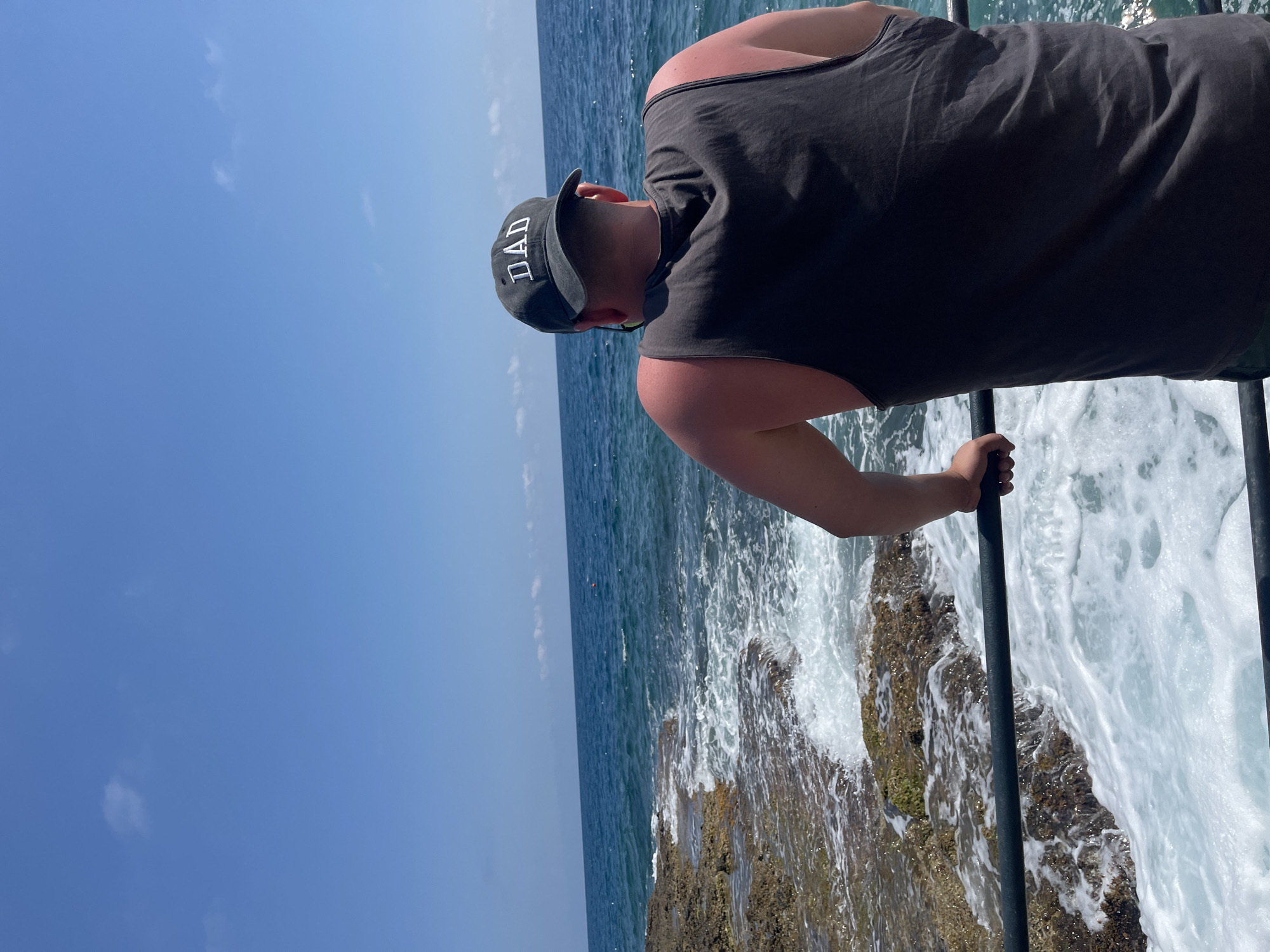 A man wearing a black tank top and a black baseball cap that says "DAD" stands with his back to the camera, leaning on a metal railing. He is facing the ocean, which is blue and has waves crashing against a rocky shore. The sky is clear and blue with a few scattered white clouds. The scene appears to be a coastal area, possibly in Alanya, Turkey, given the typical appearance of the Mediterranean coast in that region, characterized by rocky shorelines and clear blue waters. The man's shoulders and upper back are visibly sunburned. The overall impression is a peaceful, sunny day by the sea.
