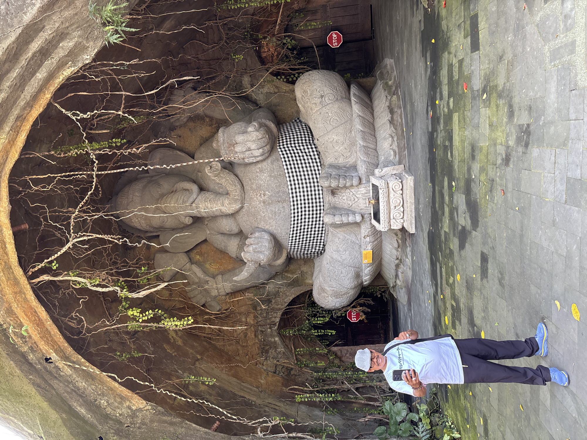 In Blahbatuh, Indonesia, a man in a white t-shirt, dark pants, and a gray hat stands in front of a large stone statue of Ganesha, positioned within a rocky alcove draped with vines. The man is holding a smartphone and appears to be taking a selfie, giving a thumbs-up with his left hand. The Ganesha statue, adorned with a black and white checkered cloth around its waist, is seated on a carved base. To the right of the statue, a red 'STOP' sign is visible on a wooden door. The ground is paved with stone tiles, some of which are damp and mossy, with scattered fallen leaves. The overall scene suggests an outdoor temple or cultural site.