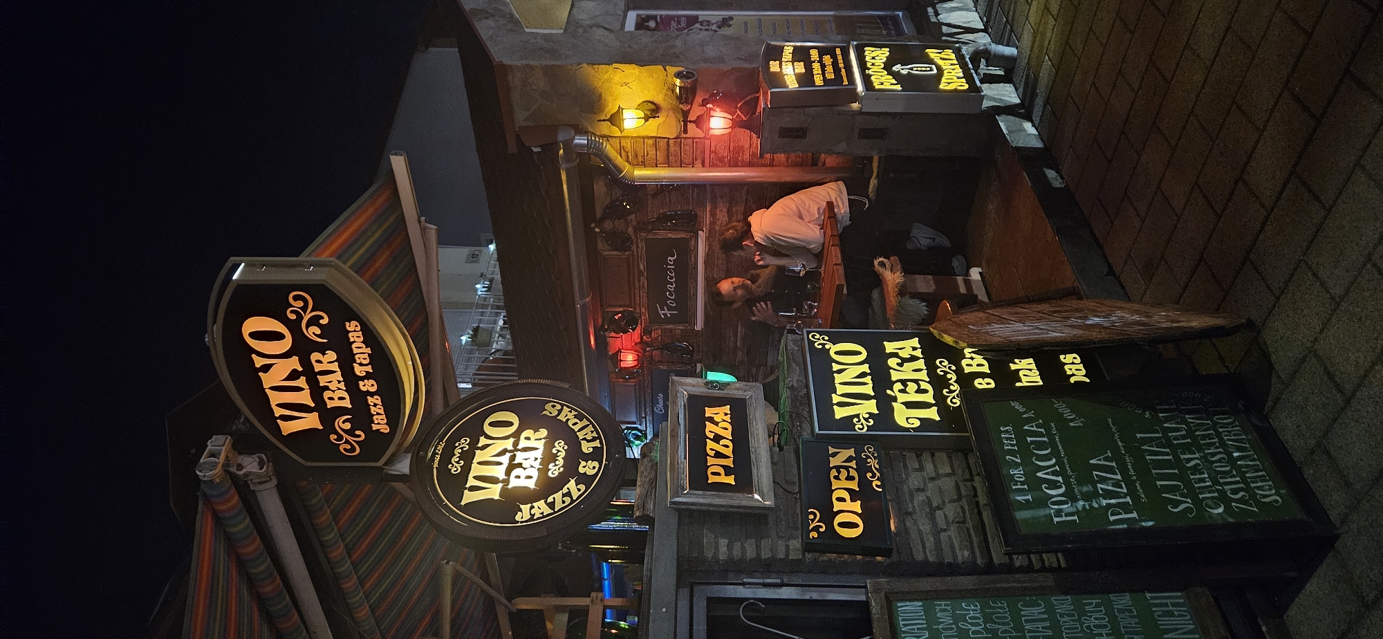 An exterior shot at night captures a lively bar and restaurant scene. The establishment, identified as "VINO BAR JAZZ & TAPAS," features illuminated signs with a vintage aesthetic, displaying the name of the bar, its offerings, and the hours of operation. A striped awning extends over the entrance, providing cover. Inside, two individuals are seated at a wooden table, engaged in conversation, with glasses of wine on the table. The building's facade is constructed of brick and stone, adorned with various decorative elements and lighting fixtures. Signs advertising "PIZZA," "Focaccia," and "FROGGZ! SPRITZ!" are prominently displayed, indicating a diverse menu. The ground is paved with cobblestones, and the overall atmosphere suggests a welcoming and vibrant nightlife setting in Hévíz, Hungary.