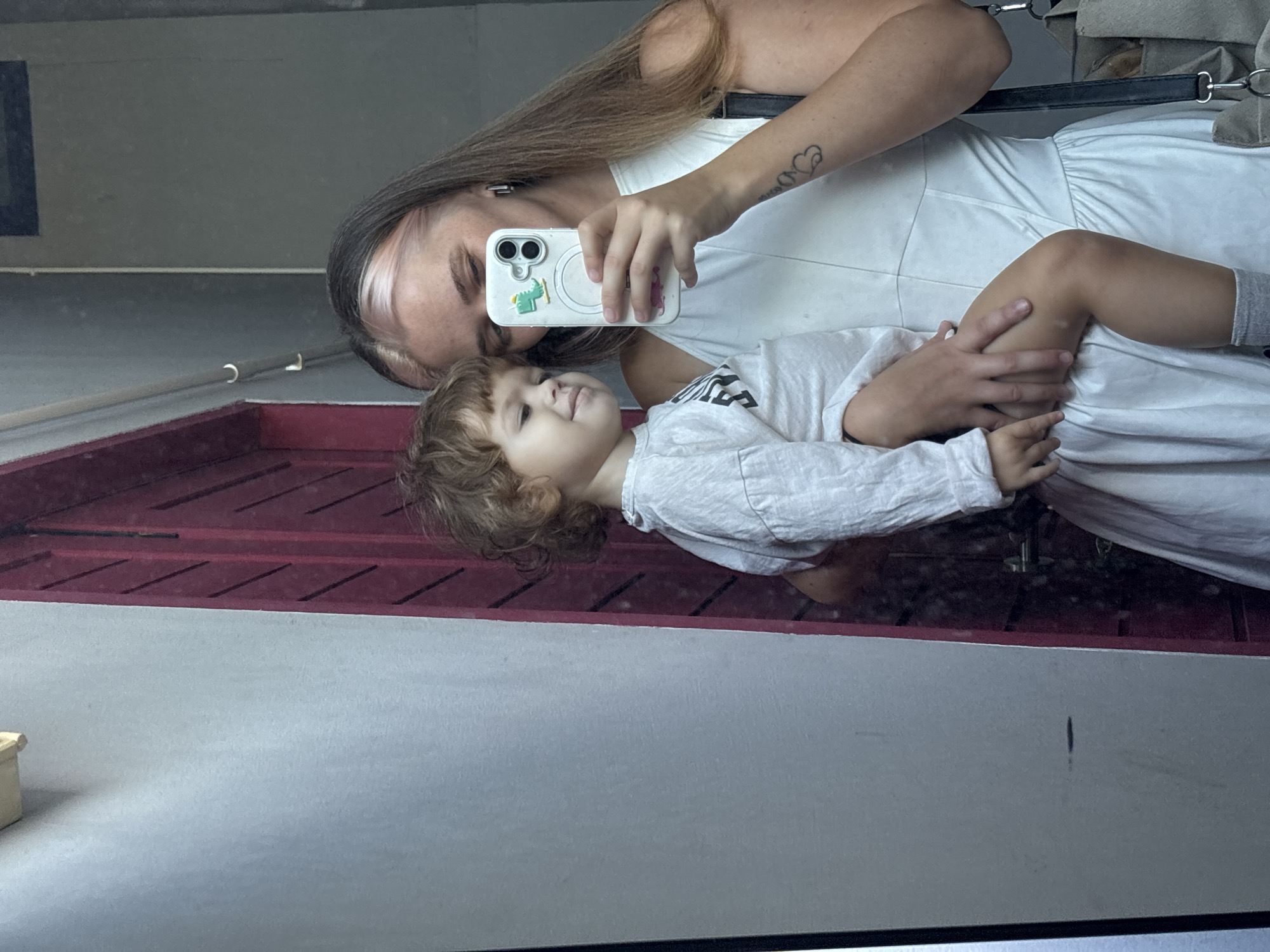 In a dimly lit urban setting, possibly a street in Choeng Thale, Thailand, a woman and a young child are captured in a mirror selfie. The woman, with dyed blonde streaks in her brown hair, holds the child, who has curly brown hair, in her arms. Both are dressed in white. The woman is wearing a white dress and is holding up a smartphone with a white case adorned with a green dinosaur sticker. The child is wearing a white long-sleeved shirt and grey socks. A red door is visible in the background, reflecting in the mirror alongside a light-colored wall. The woman has a tattoo on her left forearm. A black bag with fabric straps is slung over her shoulder. The overall mood appears casual and intimate.