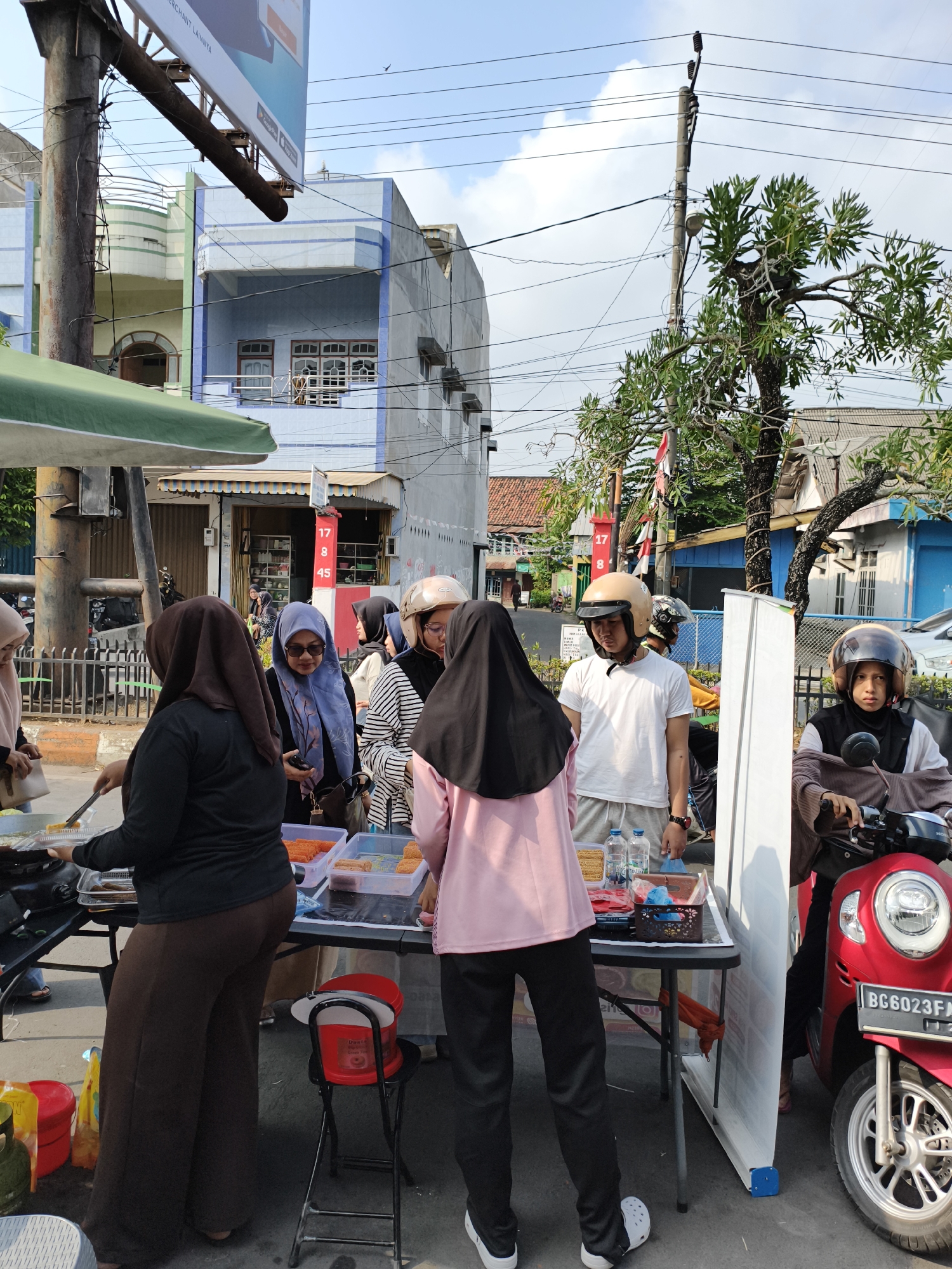 In Baturaja, Indonesia, a street scene unfolds with individuals engaged in commerce. A person wearing a black headscarf and brown trousers stands on the left, tending to a portable cooking station with a spatula in hand. To their right, a woman in a blue headscarf and sunglasses observes the items on a folding table, which displays plastic containers filled with food items, likely snacks or prepared meals. Adjacent to her, a group of individuals, some wearing helmets, appear to be customers or vendors. One person, a male wearing a white t-shirt and light-colored shorts, is facing forward, while another, a female in a pink top and black pants, is partially visible. A red scooter with license plate BG 6023 FA is parked on the right, with a person wearing a helmet and brown jacket seated on it, looking towards the front. The background reveals multi-story buildings with light blue and grey facades, indicative of urban architecture, and a clear sky with scattered clouds. Overhead, power lines and a street lamp are visible, contributing to the streetscape. The scene suggests a bustling outdoor market or food stall operation.