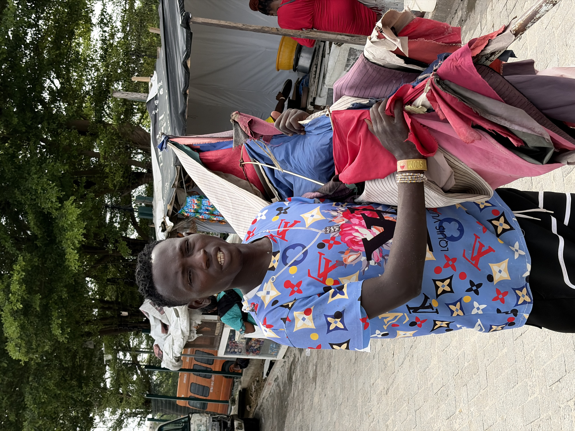 A man stands smiling, wearing a blue, short-sleeved shirt with floral and "Louis Vuitton" patterns, along a street in Igboefon, Nigeria. He's carrying a large bundle of colorful cloths over his shoulder. He wears a yellow wristband and multiple gold-colored bracelets on his left wrist. Behind him, there's a makeshift stall or tent with various items for sale, including more cloths. In the background, there are vehicles parked near the curb and trees lining the street. The environment seems to be an outdoor market area.