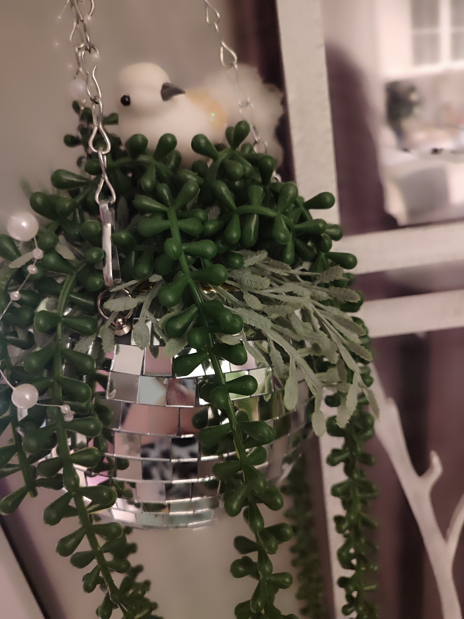 A close-up shot presents a decorative disco ball adorned with artificial succulents, suspended from a chain. The ball is composed of small mirrored squares, reflecting ambient light and nearby colors. Green succulent tendrils cascade down the sides of the ball, interspersed with silvery-gray artificial foliage. A decorative white bird is perched atop the ball, partially obscured by the plants. The hanging arrangement is placed within an interior setting, adjacent to a white shelving unit and a decorative white tree branch. The location of the scene is not discernible from the provided image. The city of Kotlas, Russia is not referenced in the image.