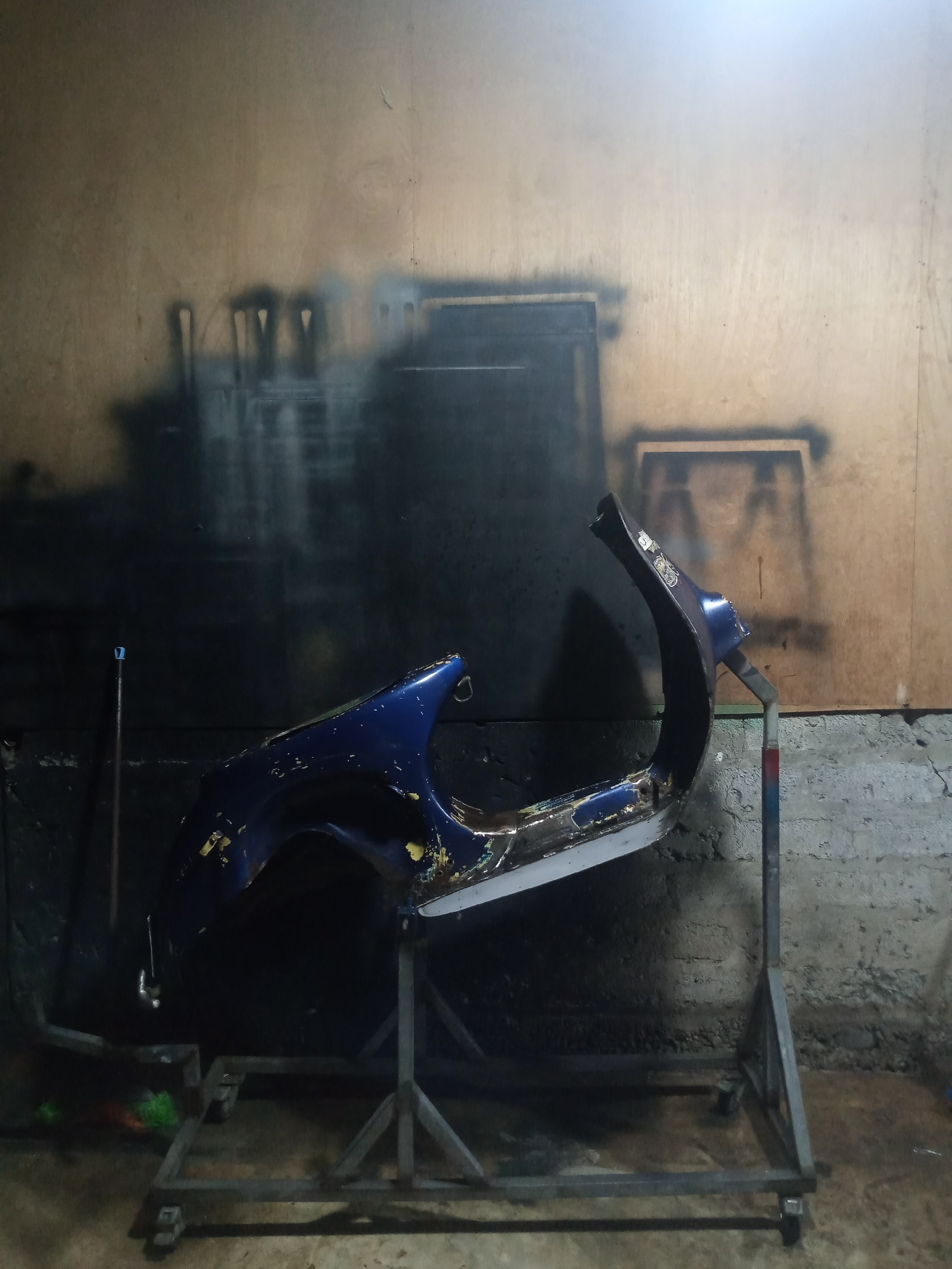The image depicts a dark, cluttered garage or workshop. In the center, a blue, damaged car fender is mounted on a metal stand. The fender shows signs of wear and tear, with chipped paint and exposed metal. The background consists of a wooden wall with spray paint marks, suggesting a work area. A broom and other tools are visible on the floor. The overall impression is one of a DIY repair or restoration project in a somewhat unkempt environment. The location is specified as Balee Rastong, Indonesia.