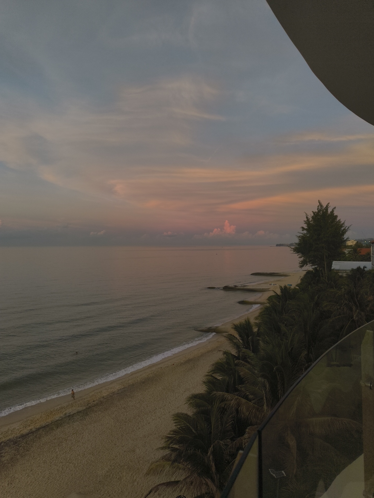 The image depicts a tranquil beach scene at dusk, likely in Phu Thuy Ward, Vietnam, with a view of the sea and a sandy shore. The sky is adorned with soft hues of pink and blue, characteristic of a sunset or sunrise. Gentle waves lap the shore, creating a serene atmosphere.
In the foreground, a balcony railing made of glass and metal frames the view. To the right, lush green palm trees line the beach, their fronds swaying gently. A single person is visible walking along the water's edge, leaving faint footprints in the wet sand.
The sea is calm, with subtle ripples reflecting the pastel colors of the sky. Small boats or buoys can be seen in the distance, suggesting some maritime activity. Rocky outcrops emerge from the water near the shore, adding to the natural beauty of the landscape.
In the background, distant buildings and a prominent tree are visible, hinting at the proximity of a town or resort. The overall impression is one of peace and natural beauty, enhanced by the soft, diffused light of the twilight hours.