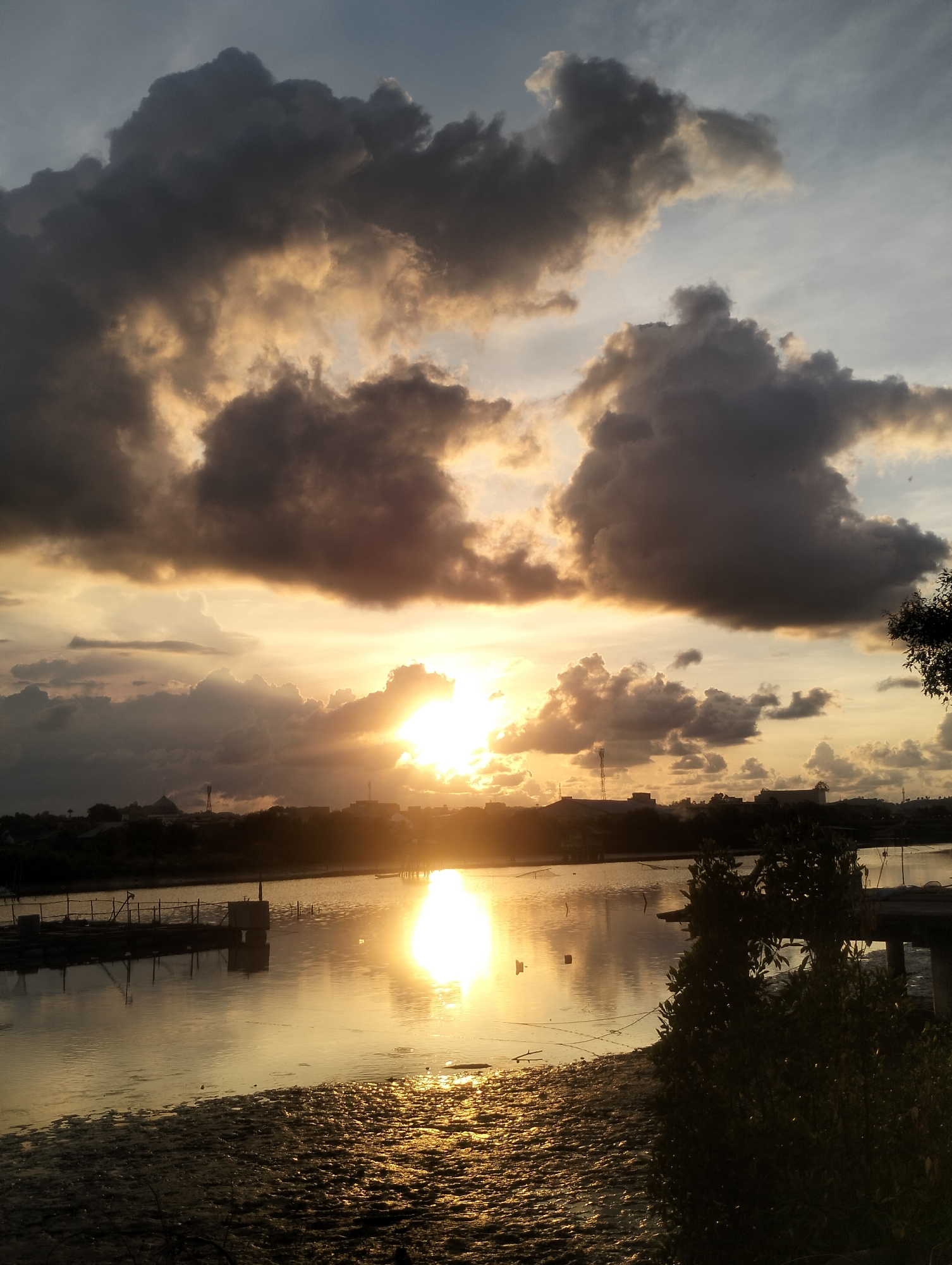 The image shows a sunset over water in Lhokseumawe, Indonesia. In the background, the silhouette of buildings and a communications tower is visible against the bright sun. The sky is filled with a mix of dark, heavy storm clouds and lighter, sunlit clouds. The water reflects the bright sunlight, creating a shimmering effect. In the foreground, there is a muddy shoreline with some vegetation growing along the edge of the water. A partially-visible wooden dock with posts extends into the water to the left.