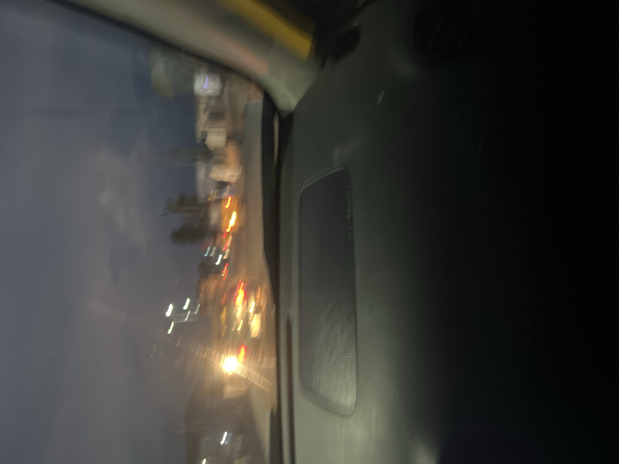 The image depicts a blurred, low-light view from the interior of a vehicle, looking out through the windshield. The scene suggests nighttime driving on a road in Jalingo, Nigeria, with car headlights and taillights creating streaks of light. The sky is dark and overcast, with some diffused light visible. The dashboard of the vehicle is partially visible in the foreground, with a textured surface. Out of focus, there are blurred shapes that appear to be buildings and streetlights along the roadside, typical of an urban environment. The overall impression is one of motion and dim visibility during a drive in Jalingo.