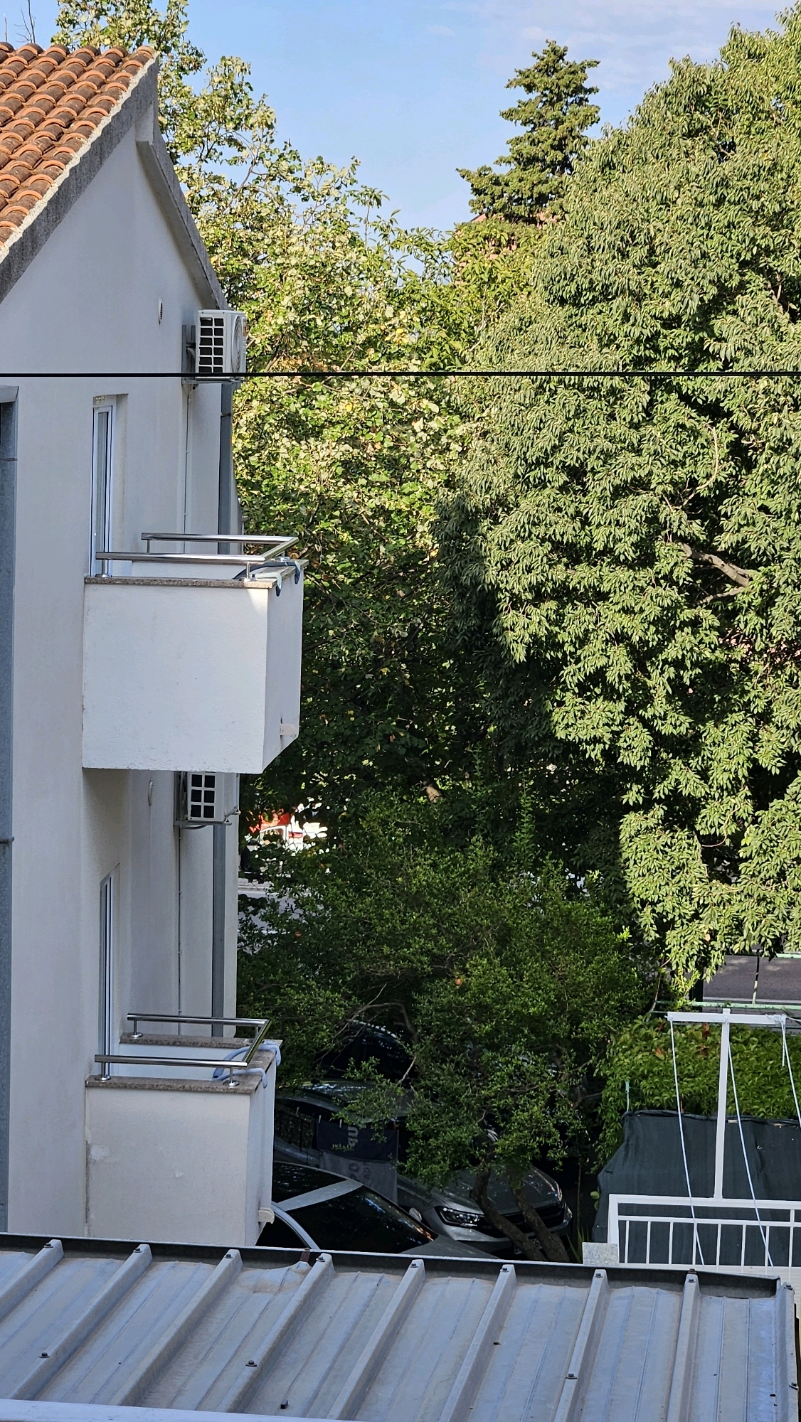 The image depicts a residential building with a tiled roof and white balconies, set against a backdrop of lush green trees. One balcony has a chrome railing and a white outer wall. An air conditioning unit is mounted on the exterior wall of the building. Below the balconies, several cars are parked, some partially obscured by foliage. In the foreground, a corrugated metal roof is visible. The sky is a clear blue. The scene is located in Općina Tučepi, Croatia.