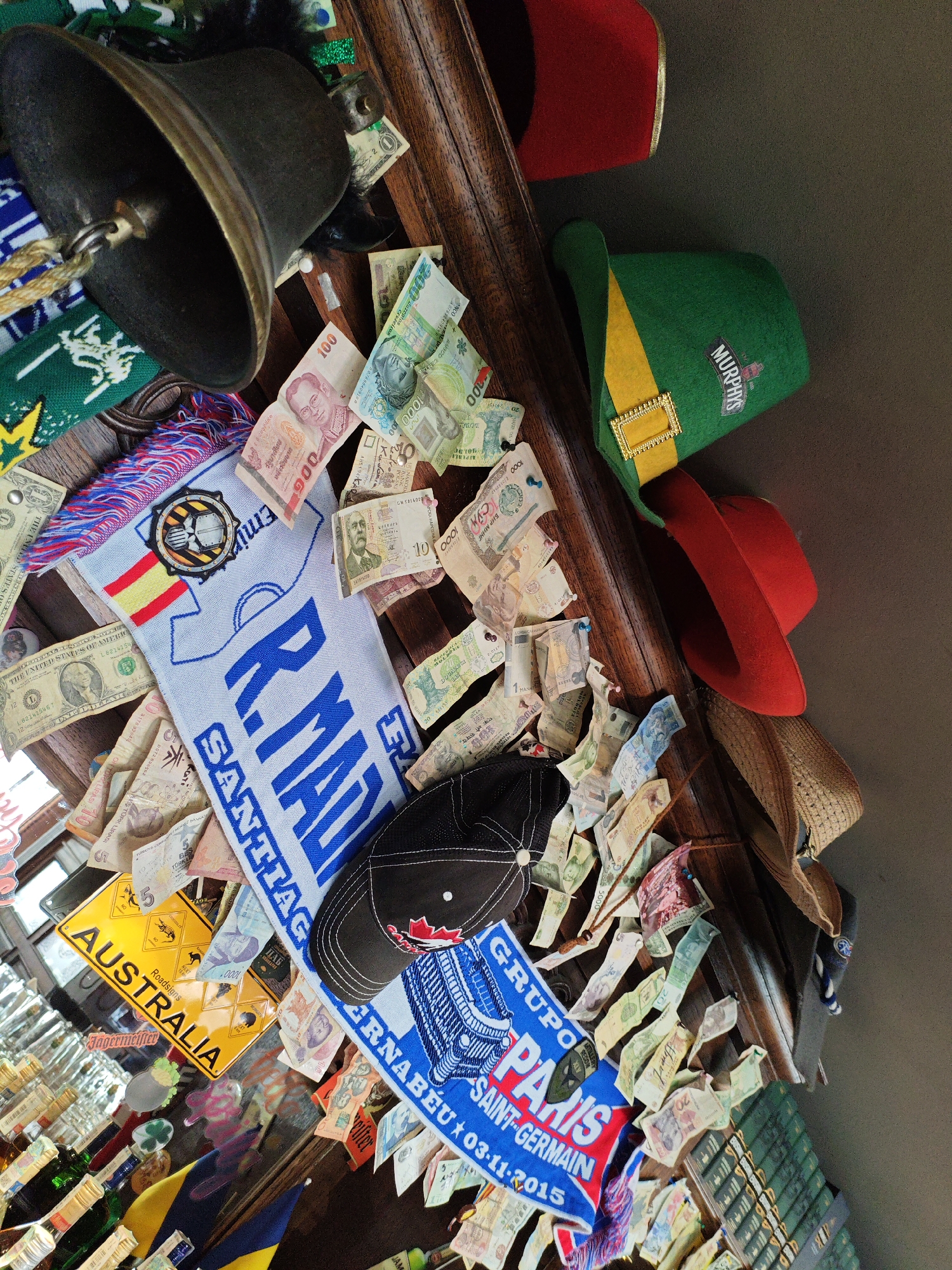 The image depicts a densely decorated shelf or bar top in what appears to be a pub or restaurant. The surface is covered with various items, primarily foreign currency notes, flags, hats, and a bell.
Specifically, there are numerous banknotes from different countries, including what looks like Thai Baht, US Dollars, and other currencies that are difficult to identify precisely due to their small size and orientation. Several hats are visible: a green leprechaun-style hat with "Murphys" embroidered on it, a red hat, a black baseball cap with a red emblem, and a woven straw hat.
A blue and white scarf with "R. MADRID SANTIAGO BERNABEU 03-11-2015" is draped across some of the items, suggesting a connection to Real Madrid football club. Another smaller scarf or textile with Spanish flag colors (red and yellow) is also present. A yellow license plate with "AUSTRALIA" and road signs is displayed. A large, dark-colored bell with a brass rim hangs from above.
The background is dark, with some suggestions of bottles and shelving on the left and right sides. The overall impression is one of a collector's display or a place where patrons have left mementos.
There is no information in the image that suggests this location is in Kyiv, Ukraine.