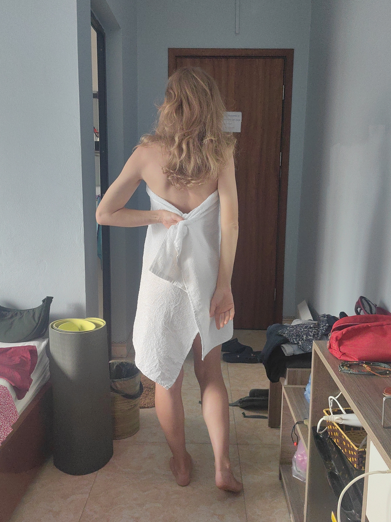 A woman with long, wavy blonde hair is standing indoors, facing away from the camera. She is partially covered by a white bath towel wrapped around her body. Her right arm is bent, and her hand appears to be adjusting or holding the towel at her back. She is barefoot, and her skin is fair.
The room seems to be a hotel room or a personal residence. A bed with scattered items is visible on the left. In the background, a wooden door occupies the center. To the right, a wooden shelving unit with various items is positioned. Scattered clothes and a bag are visible on the floor. The walls are painted light blue, and the floor is tiled with a light-colored, patterned tile.
Given the information and surroundings, it is probable that this image was taken at accommodation of some kind, either a hotel or Airbnb in Bai Chay Ward, Vietnam, or at a property in a similar style.