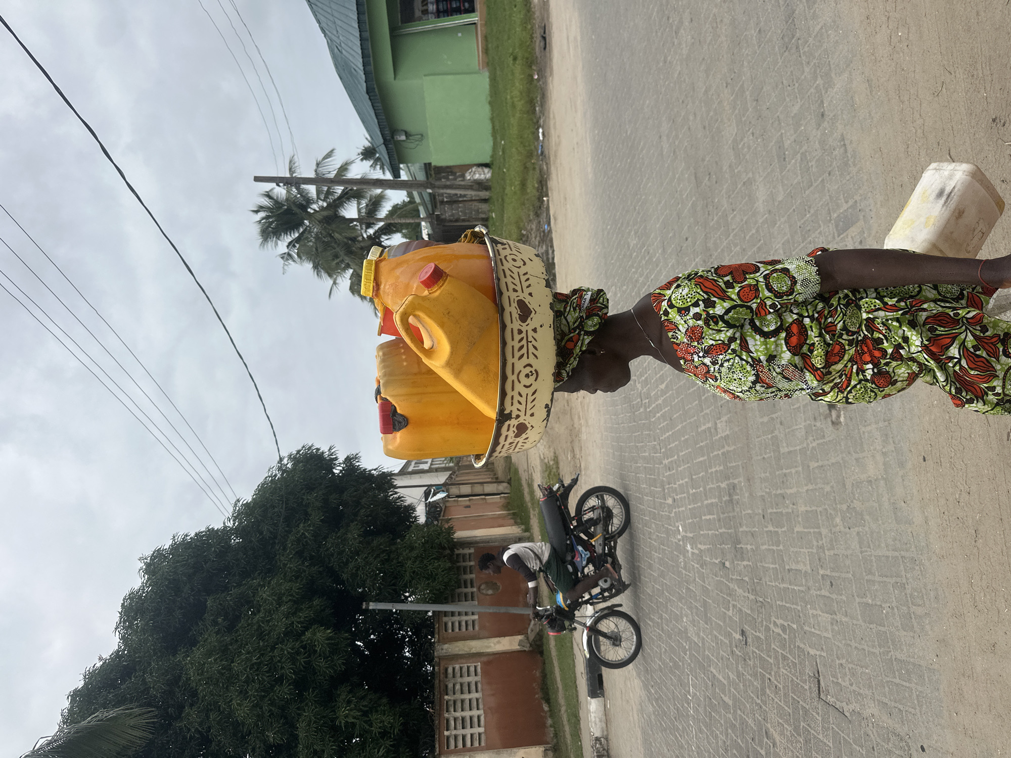 An outdoor, medium shot shows a woman in the foreground, in profile, carrying a large metal basin filled with yellow plastic containers on her head. She is wearing a brightly colored green, red, and orange patterned dress and a matching headscarf. Her arms are down by her sides, and she is walking away from the viewer. In the mid-ground, a man is riding a black motorcycle on a paved road. He is wearing a white t-shirt and dark shorts, and is looking forward. To the left of the road is a red and white brick wall and a large, leafy green tree. In the background, there are several buildings, including a green building with a dark green roof. Palm trees are visible in the distance. The sky is overcast. This scene is set in the city of Badagry, Nigeria.
