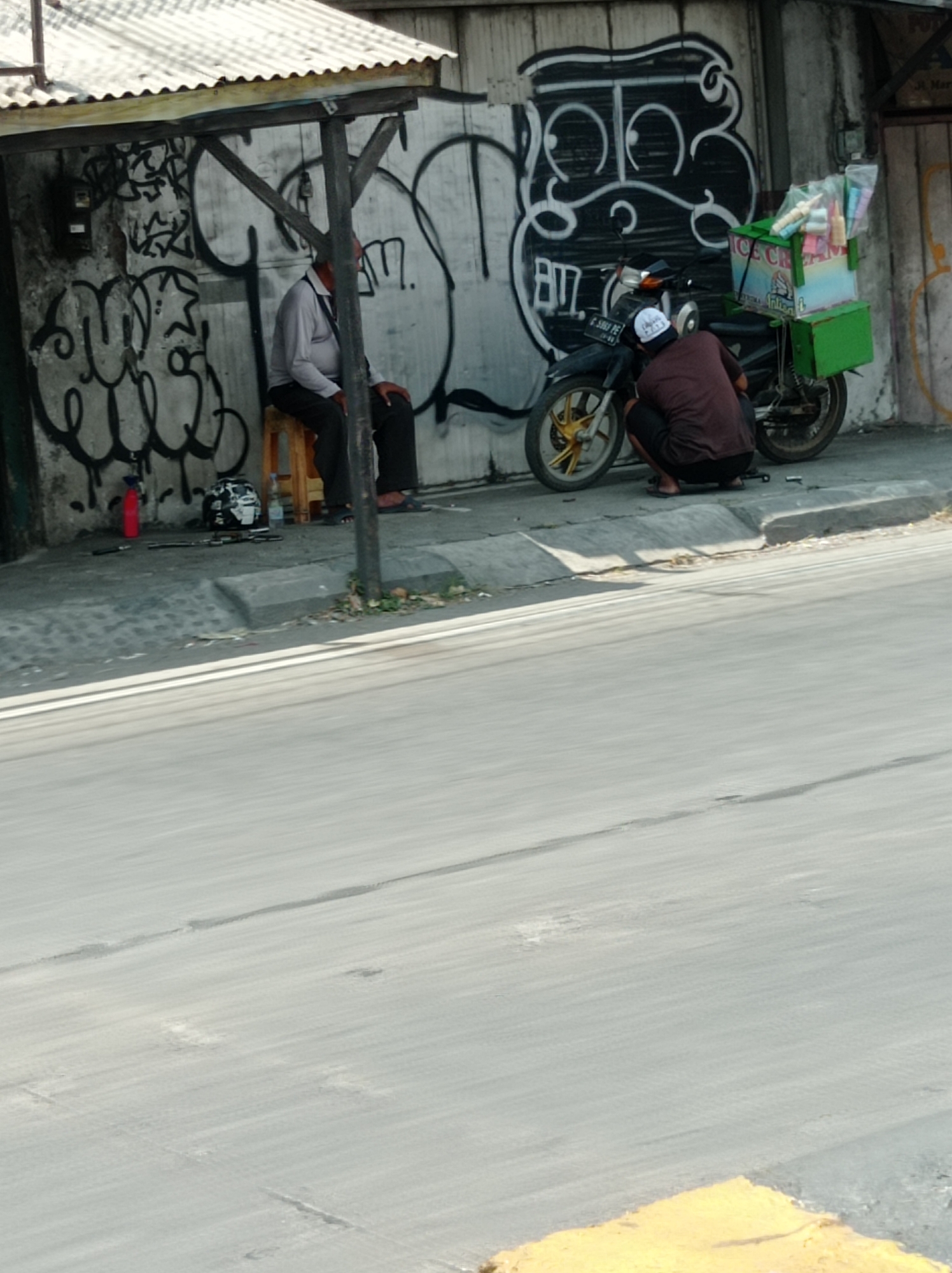The image depicts a scene in Tegal, Indonesia, likely on a street. In the foreground, a blurred asphalt road occupies a significant portion of the frame. To the right, a yellow painted curb is visible. The midground shows a sidewalk area with a man sitting on a wooden stool under a simple shelter with a corrugated metal roof. He is wearing a light-colored long-sleeved shirt and dark pants. Next to him, a motorcycle is parked, equipped with an ice cream cart. Another person, wearing a dark shirt and dark shorts, is crouched down near the front wheel of the motorcycle, possibly attending to it. The background features a concrete wall covered in graffiti, with various abstract and figurative drawings in black and white. The overall impression is of a casual street scene, possibly a vendor's spot in the city.