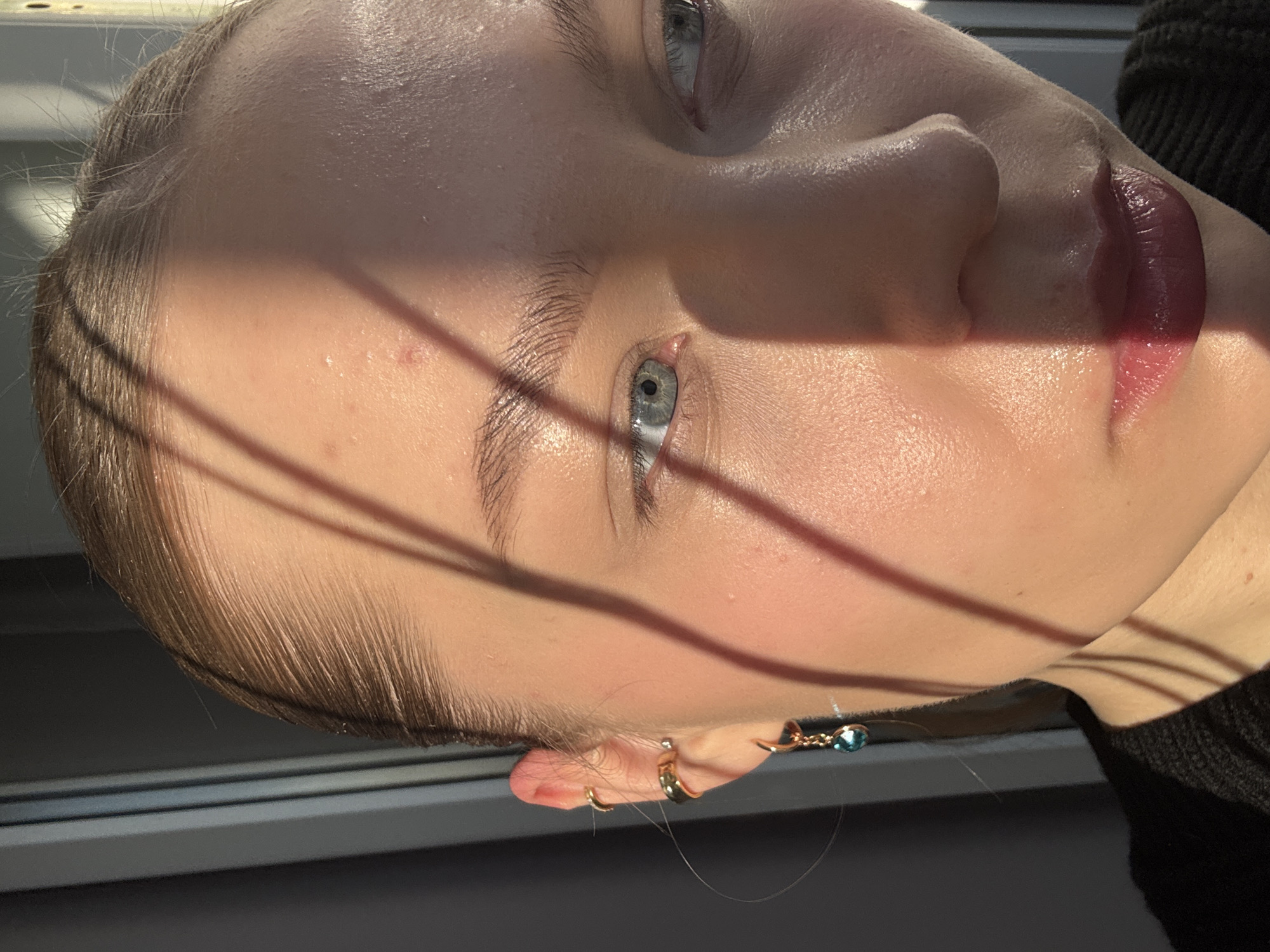 A close-up, rotated view of a person's face, predominantly illuminated on the forehead, eye, nose and lip. The person appears to have light skin, fair hair, and light-colored eyes, all partially cast in shadow. The lips are colored a deep, reddish-purple. There are multiple gold earrings in the ear. The person is wearing a dark, possibly black, garment. The setting is unclear, the location in Chaikovsky, Russia is not determinable from the image.
