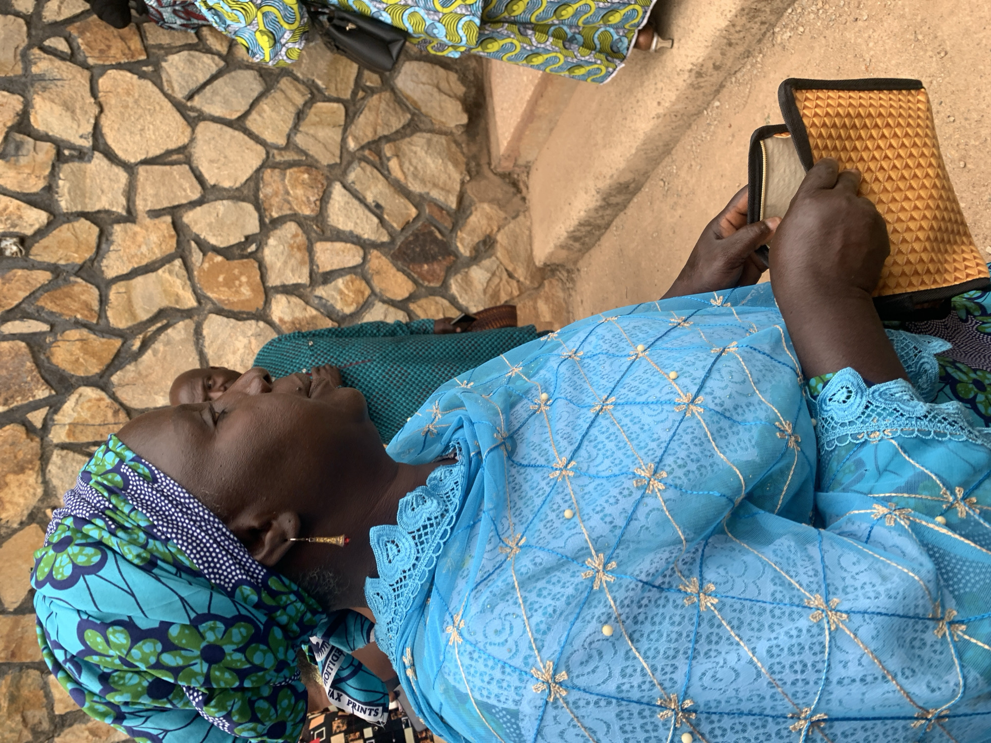 An older Black woman wearing a turquoise and blue headscarf and a turquoise lace top is sitting outside in Jos, Nigeria. She is holding a small, tan-colored zippered bag and appears to be looking inside it. The ground is made of tan stone tiles. Another person wearing a patterned cloth is partially visible in the background.
