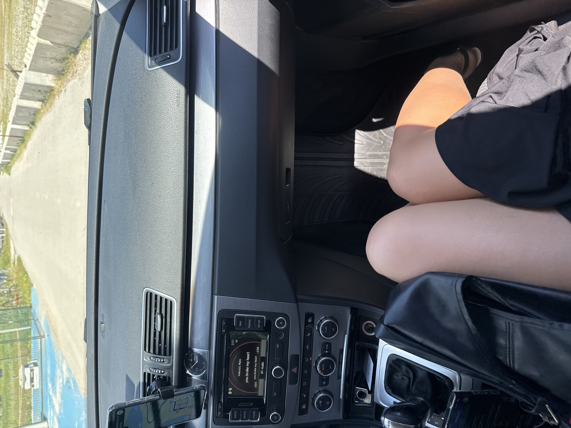 The image depicts the interior of a car, possibly a Volkswagen, with a view of a road outside the front window, located in the city of Prijevor, Montenegro. The dashboard is visible, including the air vents, clock, radio, and gear shift, along with a phone mounted to the dashboard for navigation purposes. The radio is tuned to a song called "you broke my heart." A person is sitting in the passenger seat, their legs are visible, wearing a black dress, with a black purse sitting in their lap. The image is well-lit and appears to have been taken during the daytime.
