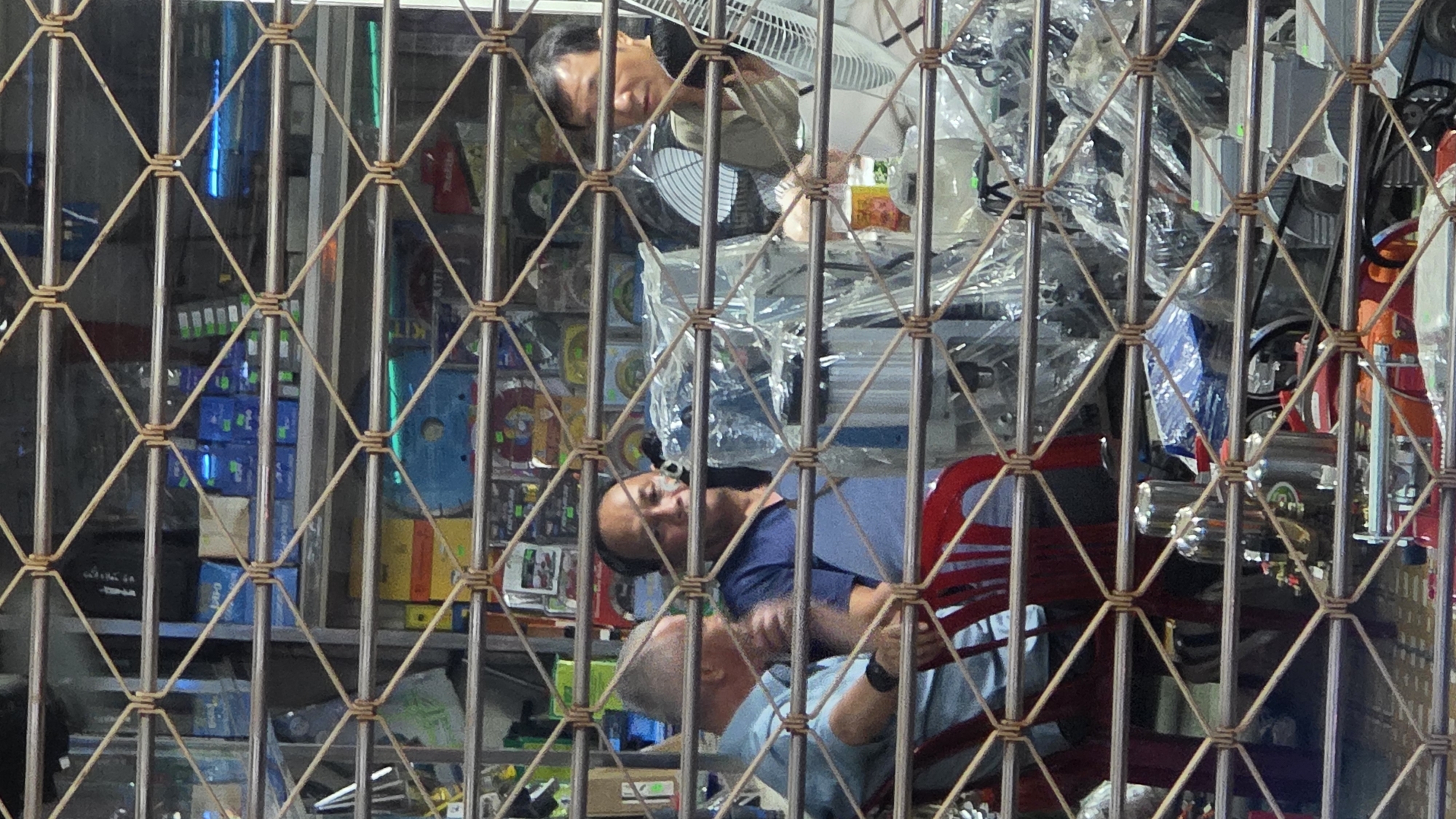The image shows the interior of a shop in Nha Trang Ward, Vietnam, as viewed through a metal security gate. Three men are visible within the shop. One man is at the top, looking down. Two men are sitting and face the gate. The shop interior is packed with merchandise, including boxes, fans, and other mechanical items, some wrapped in plastic.