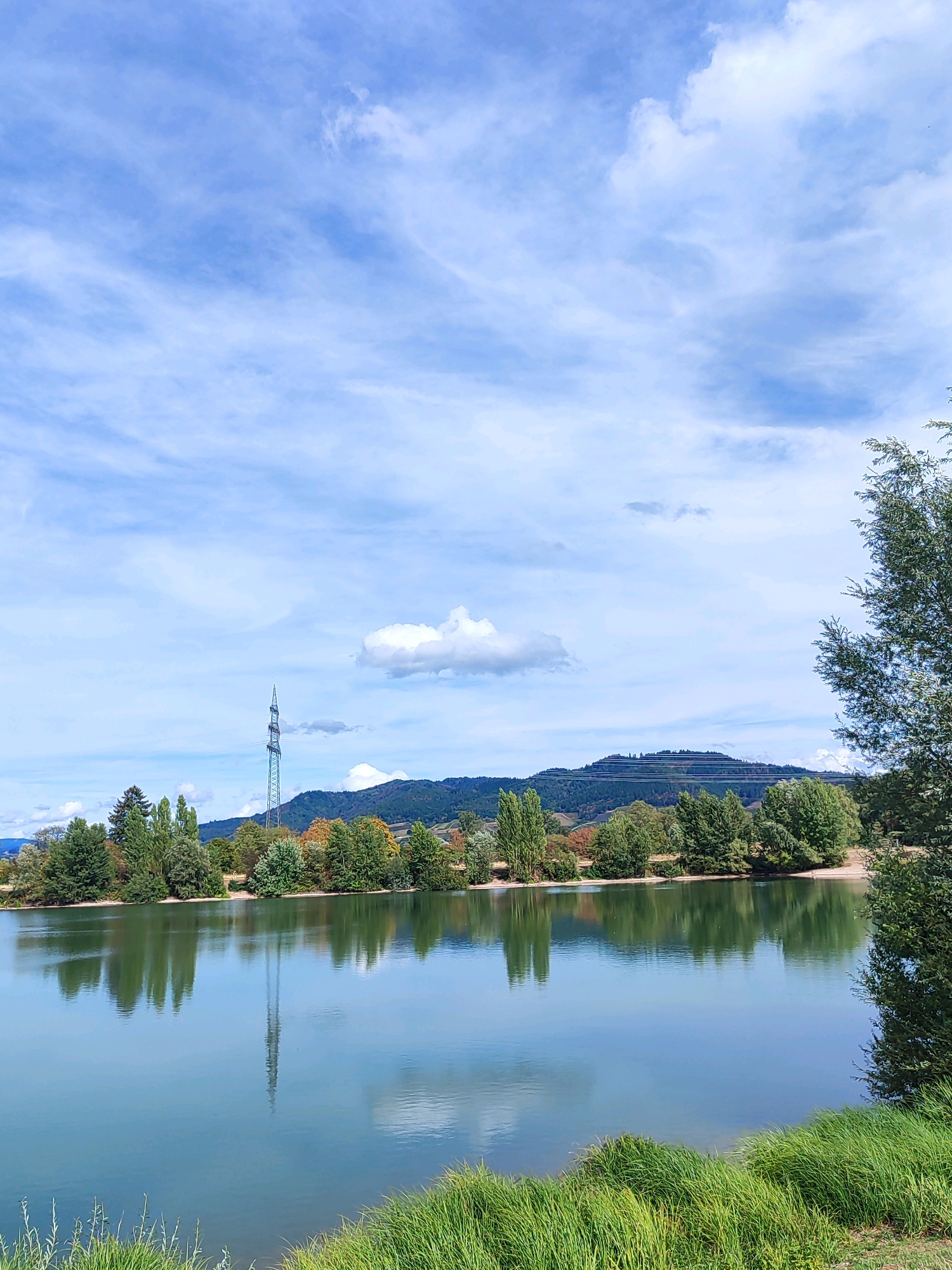 This image captures a serene landscape scene, likely near Offenburg, Germany, with a focus on a calm body of water reflecting the sky and surrounding environment. The sky is a bright blue with wispy white clouds, suggesting fair weather. In the background, rolling hills are visible, with a prominent cell tower standing tall on one of them, indicating a developed, yet still natural, area. A line of trees, some displaying autumn colors, lines the opposite bank of the water, their reflections clearly visible on the water's surface. In the foreground, lush green grass grows along the edge of the water, adding to the natural beauty of the scene. There are no people or animals visible in the image, and no apparent actions or interactions are taking place. The overall impression is one of tranquility and natural beauty.