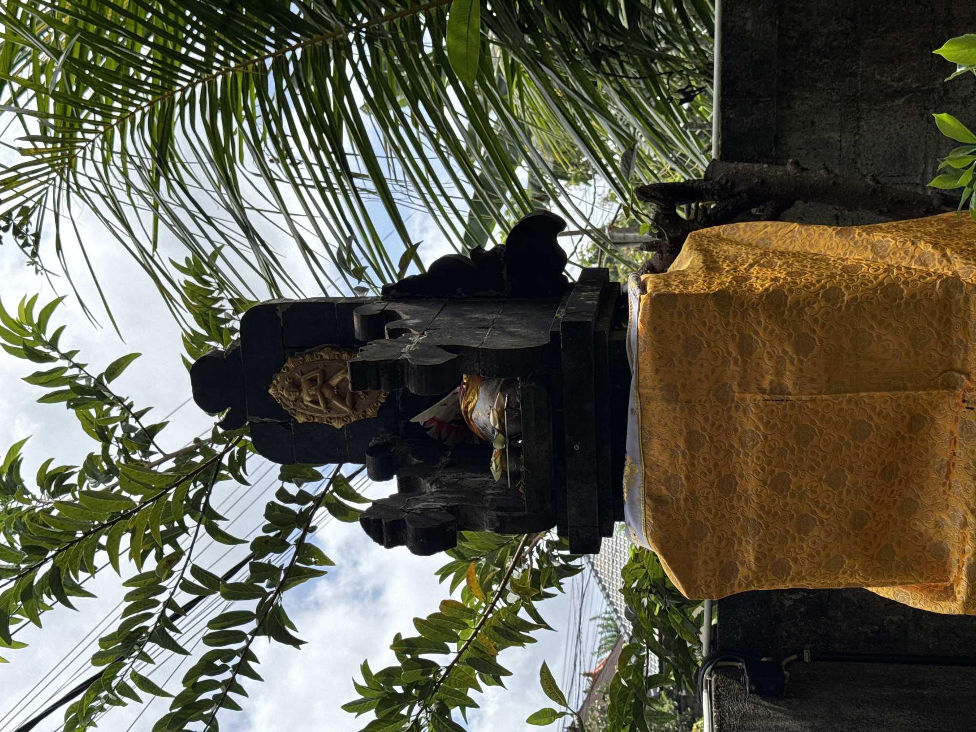 A low-angle shot shows a small, ornate black structure on a stone pillar, wrapped in a gold cloth, set against a backdrop of foliage and a partly cloudy sky. The structure features intricate carvings and a gold-colored decorative element. Green leaves and branches fill the foreground, partially obscuring the view of the sky, while palm fronds are visible at the top of the frame. The location is Kuta Selatan, Indonesia.