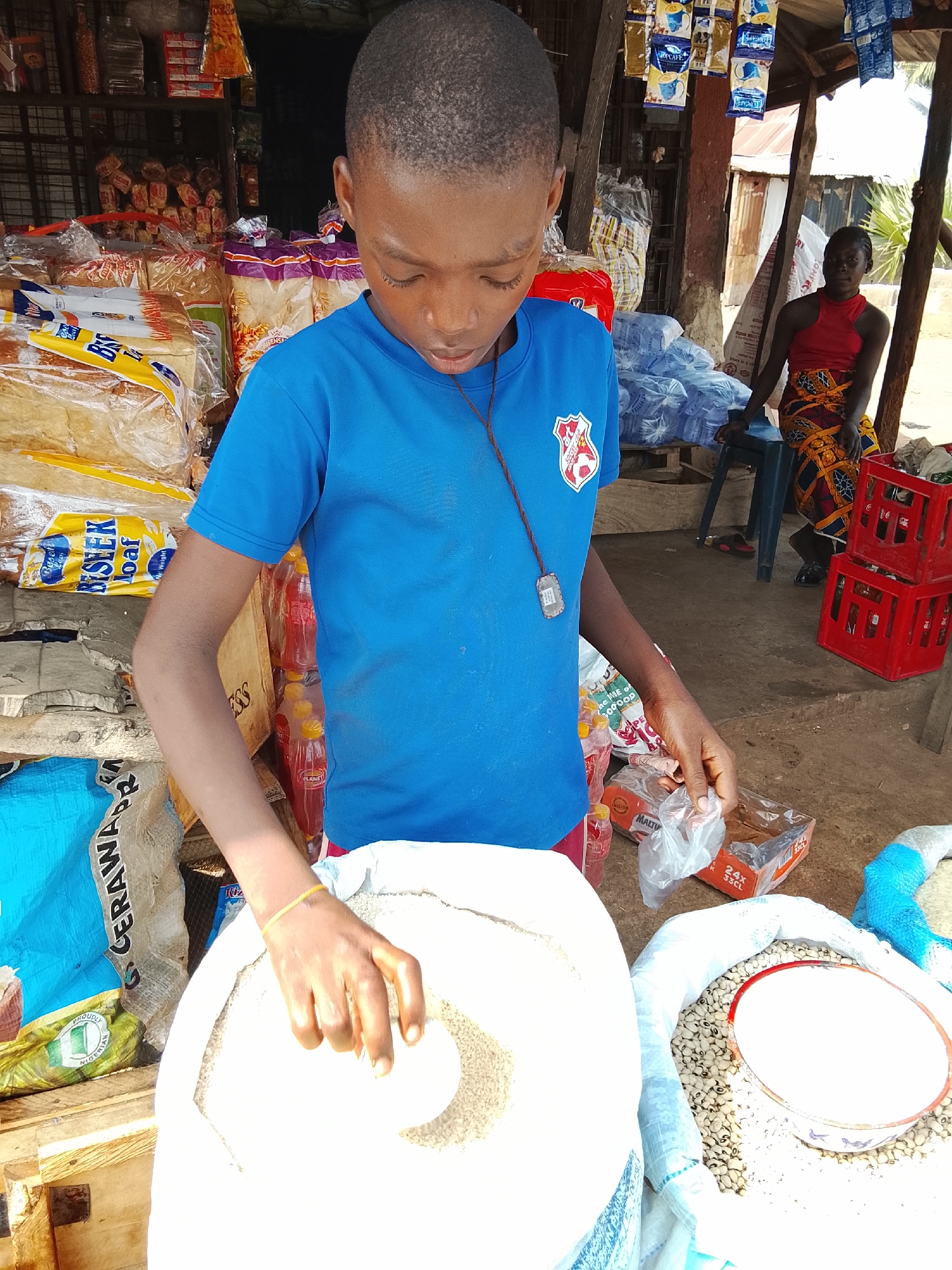 The image captures a market scene in Akwanga, Nigeria. A young person wearing a blue shirt with a school emblem is shown scooping grains from a large sack. There are other sacks of grains nearby, including one containing beans with a white bowl on top. The young person is also holding a small, clear plastic bag. In the background, a woman is seated on a blue plastic stool next to red plastic crates. The market stall is stocked with various goods, including bread, bottled drinks, and other packaged items. There are coffee packages hanging from the stall's structure.