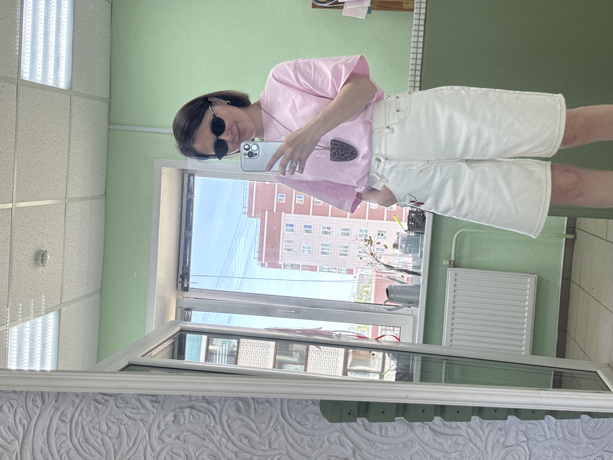 A woman takes a mirror selfie in a room with green walls and a white tile ceiling. She wears sunglasses, a pink t-shirt, and white shorts. She holds a smartphone in her left hand, and a necklace hangs around her neck. A building is visible outside the window, located in Izhevsk, Russia. A radiator is attached to the wall near the window. The mirror frame is white with a decorative pattern.