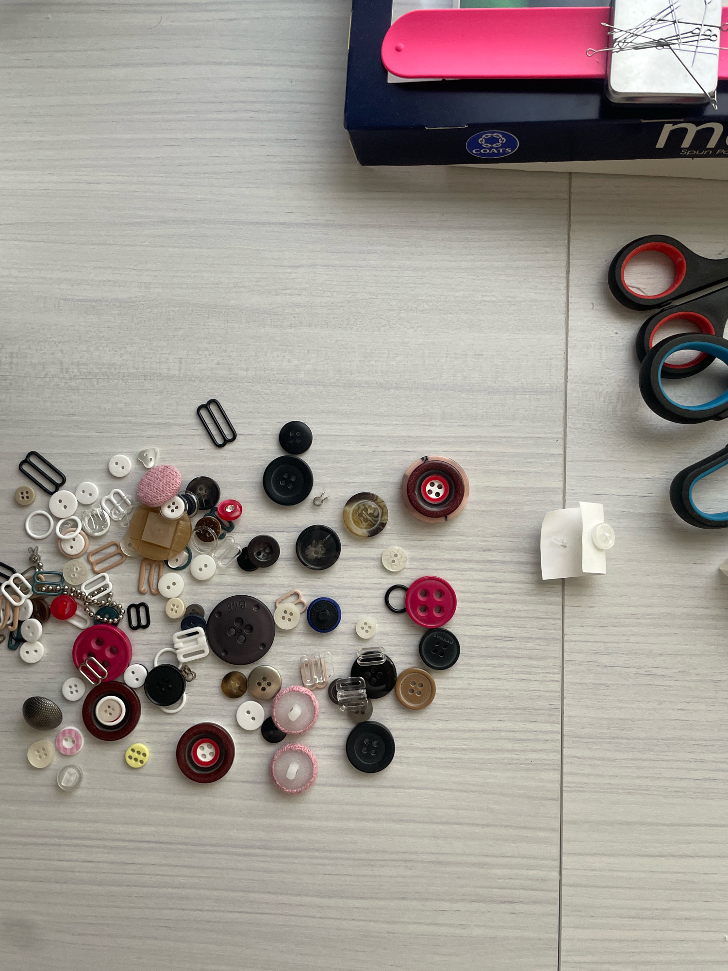 A flat lay image captures various sewing notions scattered on a light gray wooden surface. In the upper right, a dark blue box with "COATS" and "m spun pol" branding is visible. Adjacent to the box, to the right, are two pairs of black handled scissors with red and blue accents, positioned vertically. The majority of the image is occupied by a collection of buttons of various sizes, shapes, and colors, including black, white, pink, red, brown, and clear plastic. There are also some metal buckles, sliders, and a thimble. A small piece of white paper with a single white button attached is located in the middle right of the frame. The overall scene depicts sewing supplies. The image does not provide any information or context to determine a location in Atarfe, Spain.