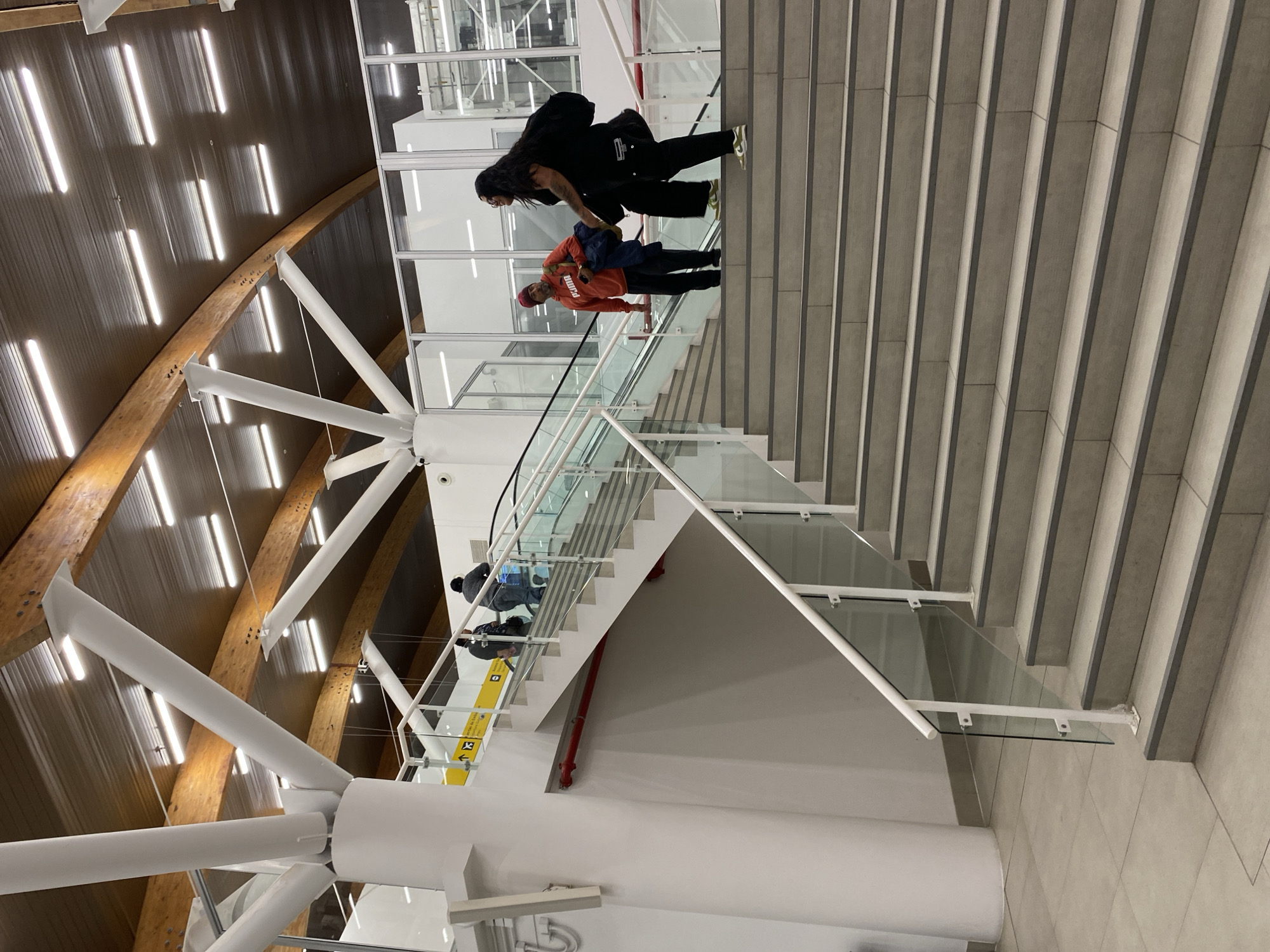 The image depicts the interior of an airport or transit hub in Arica, Chile, with architectural elements including a curved roof supported by white metal structures and a multi-level staircase with glass railings. Two individuals, a man wearing an orange Puma sweatshirt and a red hat and a woman in black clothing, are walking on the staircase, presumably going up. Other people can be seen indistinctly further down the stairs. The flooring is tiled, and overhead lighting is visible. The overall environment is modern and brightly lit.