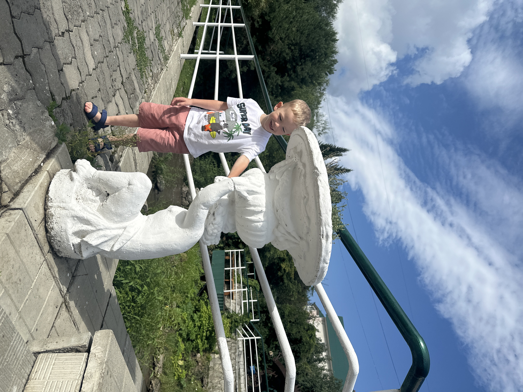 A young boy with blond hair and wearing a white t-shirt and pink shorts stands on a paved walkway next to a white, weathered statue of a cherub holding a decorative bowl. The boy is smiling and looking to his right. Behind him is a white railing. Beyond the railing, there is a body of water and lush green trees. In the background, a white building with a green roof is visible amongst the trees. The sky is blue with scattered white clouds. The scene appears to be outdoors in a park-like setting. The location is identified as Danilovka, Altai Krai, Russia.