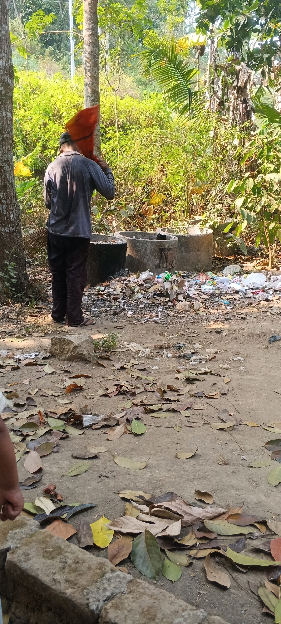 A person in dark pants and a grey long-sleeved shirt is standing in an outdoor area with scattered trash and fallen leaves. The person is holding a bright orange object over their head with their left hand and a broom with their right hand, suggesting they are engaged in cleaning or carrying something. The background is filled with dense green foliage, indicating a natural or semi-natural environment. Two large concrete circular structures are visible, one appearing to be a pit or well, and the other is filled with debris. The ground is dirt and covered with dry leaves. The scene appears to be a rural or less developed area, potentially on the outskirts of Penanggungan, Indonesia, given the lush vegetation and the visible waste accumulation.