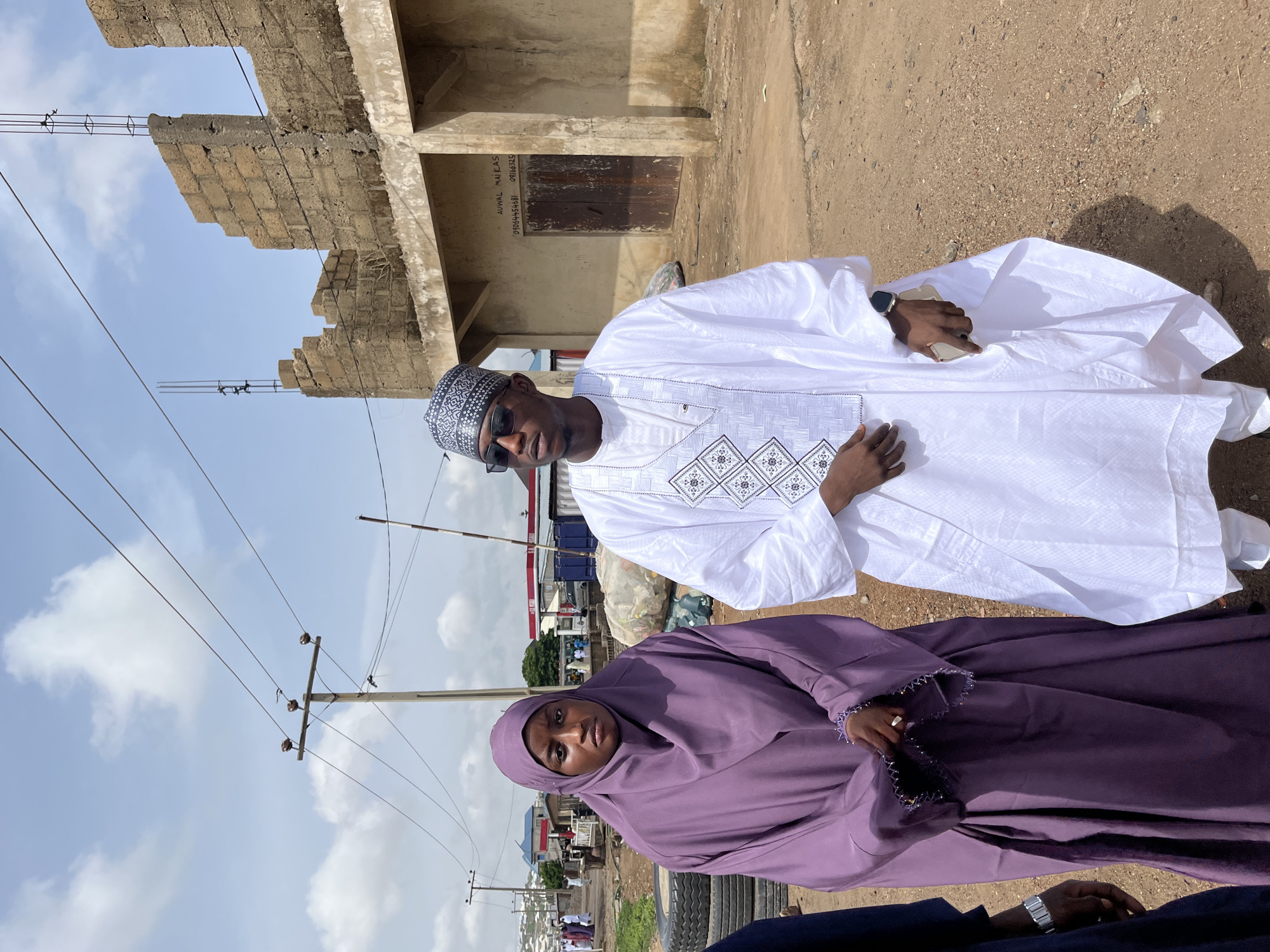 This outdoor, full-length portrait features a man and a woman posing under a bright, partly cloudy sky in what appears to be a dusty, semi-urban environment in Naraguta Hausawa, Nigeria.
The man, standing centrally, wears a traditional white *boubou* or *kaftan* with intricate grey and white embroidered patterns on the chest. He sports a patterned black and white *kufi* cap and dark sunglasses. His left wrist has a visible smartwatch, and he holds a light-colored smartphone in his right hand. He looks directly at the camera with a neutral expression.
To his left and slightly in the foreground, a woman is seen, possibly sitting or kneeling. She is dressed in a modest purple *abaya* and matching *hijab*, covering her head and body. Her right hand, adorned with a gold ring, rests on her lap and holds a string of prayer beads (tasbih). She also looks directly at the camera with a neutral expression. The arm of a third person, wearing a silver-toned watch, is visible at the bottom of the frame, beside the woman.
The background features a partially constructed or dilapidated building made of concrete blocks, with exposed rebar. On one wall of this building, faint white text is visible, reading "AUWAL MAI KAS" followed by two phone numbers: "07034454581" and "091363219". Overhead, power lines run across the scene, supported by utility poles. The ground is bare and appears dusty or earthen. The bright sunlight suggests it is daytime, possibly mid-morning or afternoon.